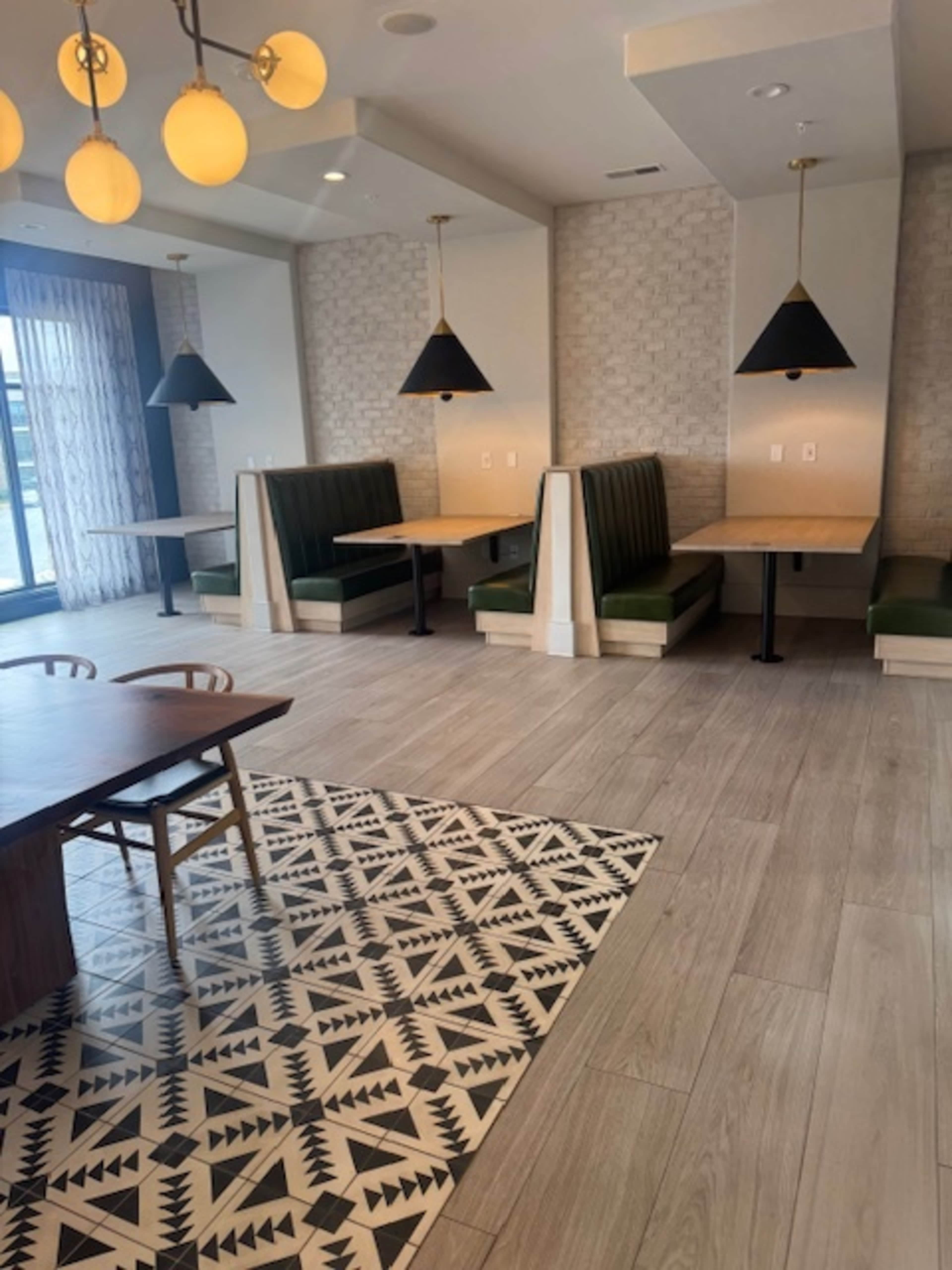The image shows a modern dining area featuring wooden tables, green booth seating, and patterned rugs under pendant lighting.