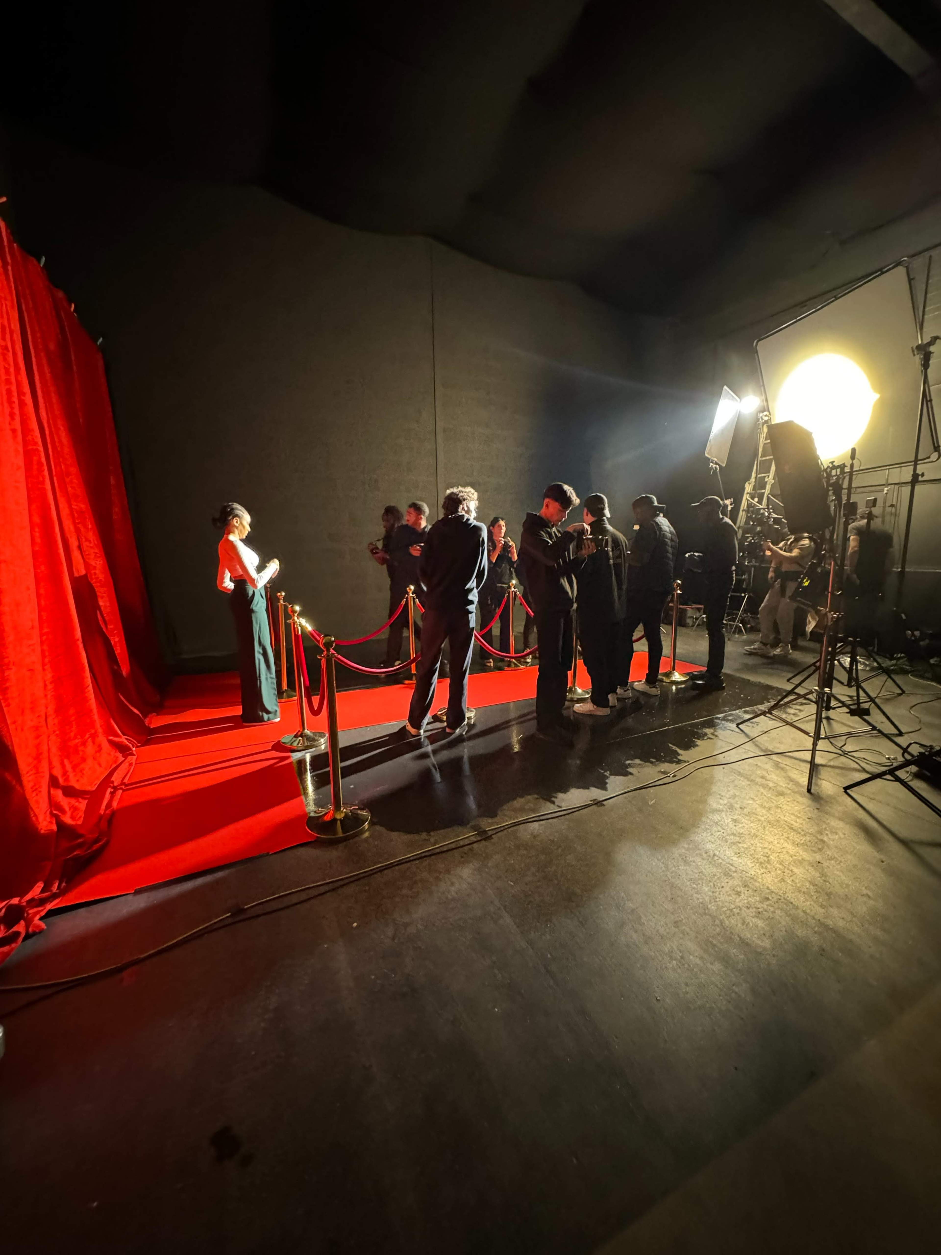 A group of people is gathered around a red carpet area with a red backdrop, where a woman stands posing for photos under bright lights.