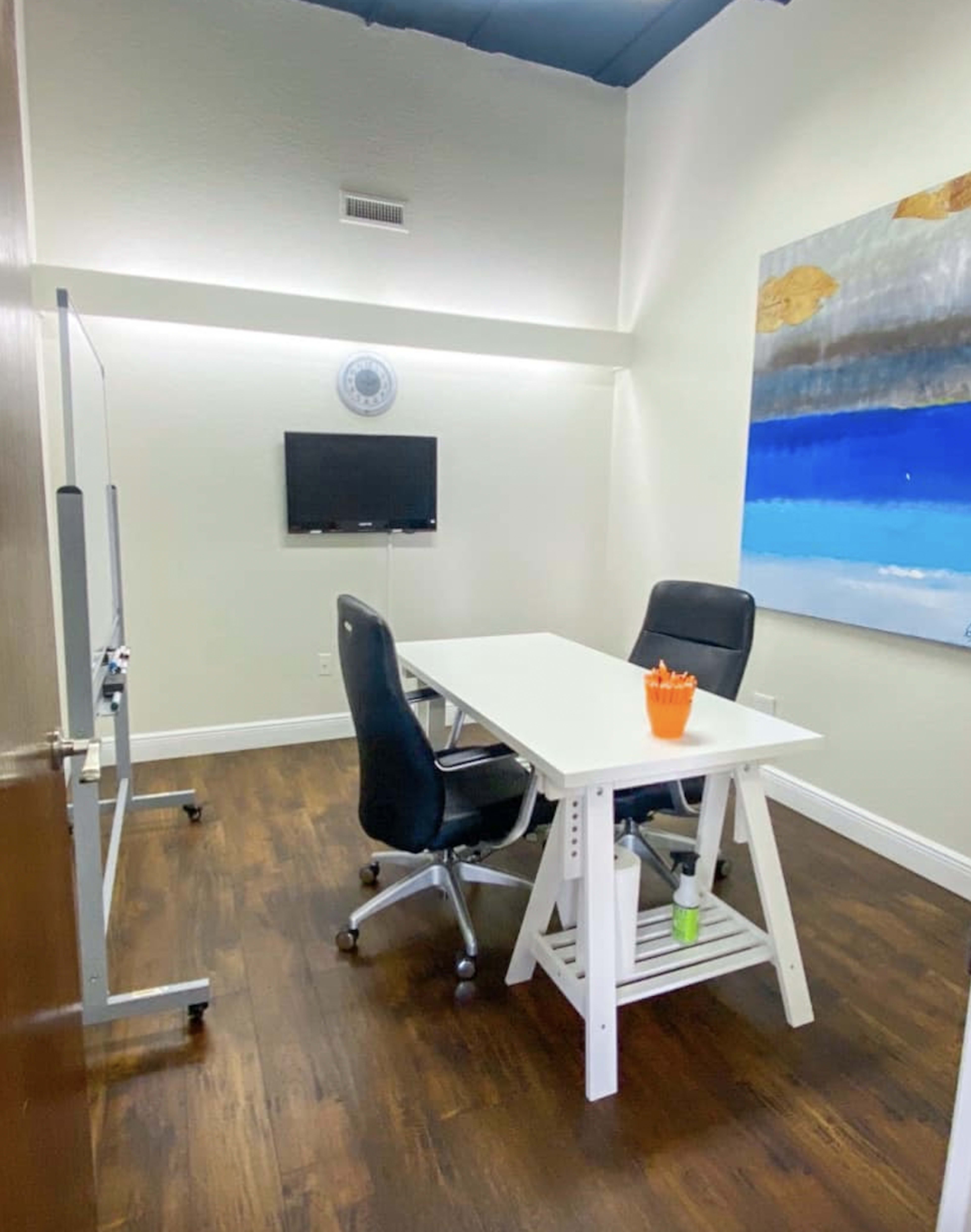 A small office featuring a white table, two black chairs, a wall-mounted TV, and a colorful abstract painting.