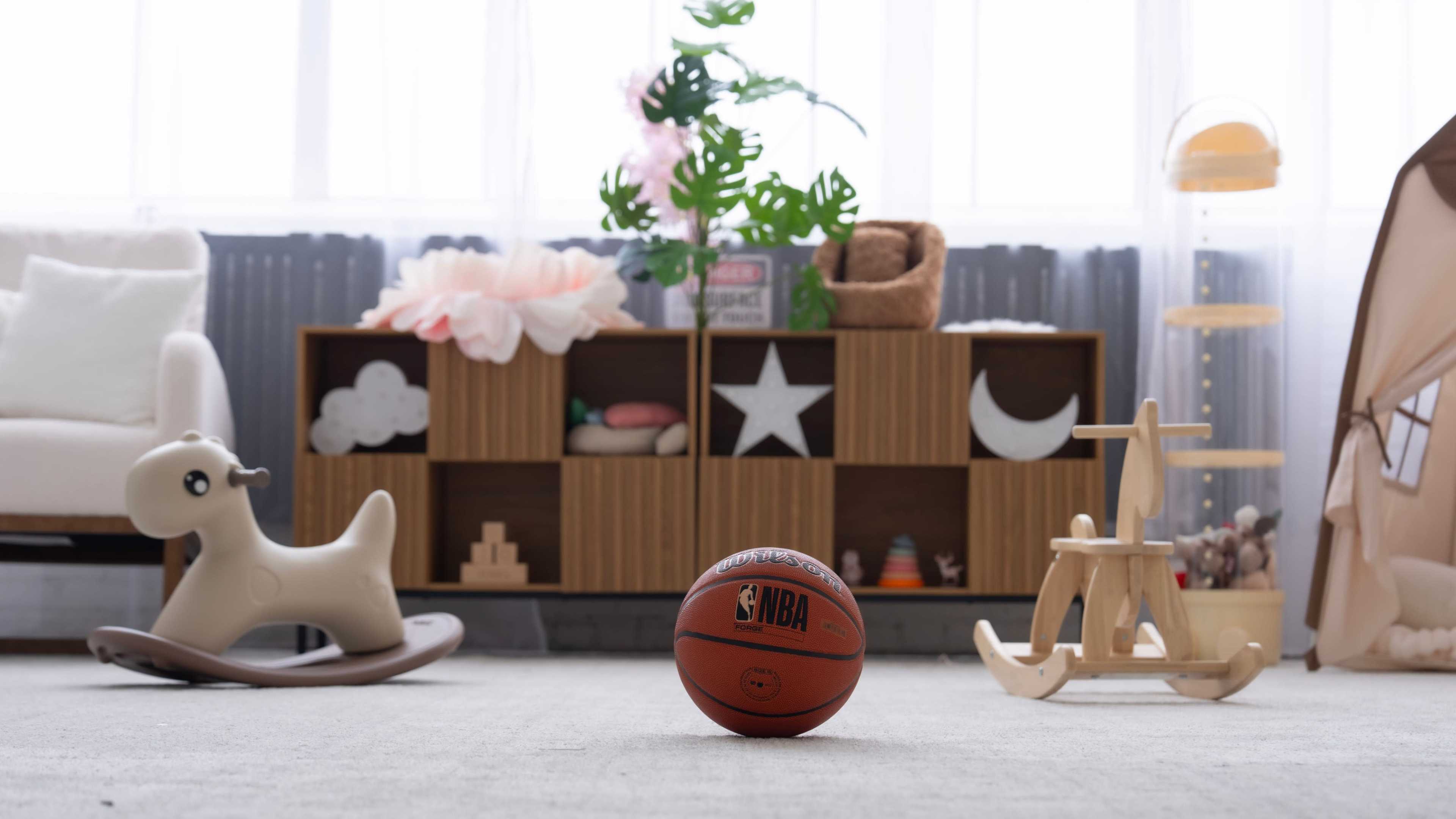 A basketball sits on a carpeted floor in a room decorated with toys, a bookshelf, and soft furnishings.