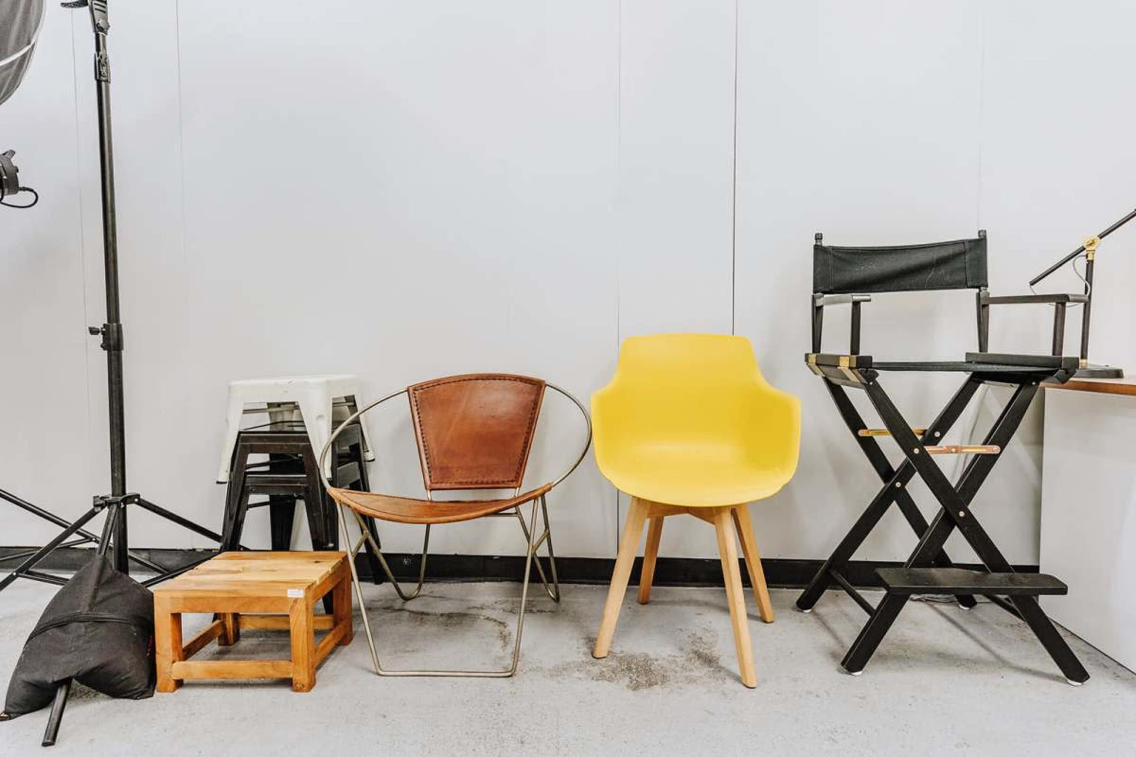 The image shows a variety of chairs, including a yellow armchair, a wooden stool, a metal chair, and a black director's chair, arranged against a plain wall.