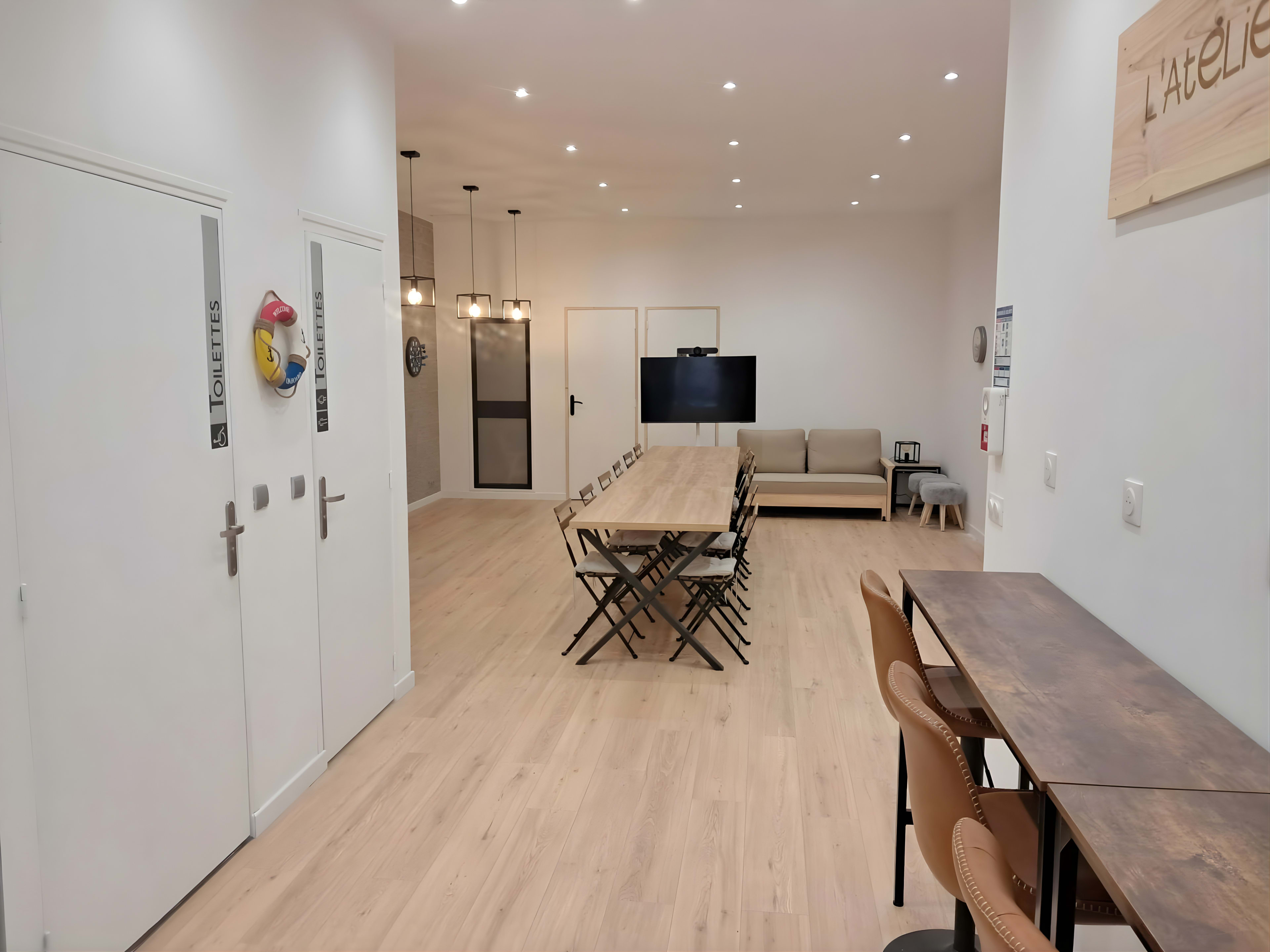 The image shows a spacious, well-lit room with a long table at its center, surrounded by chairs, and a seating area with a couch and a TV on the wall.