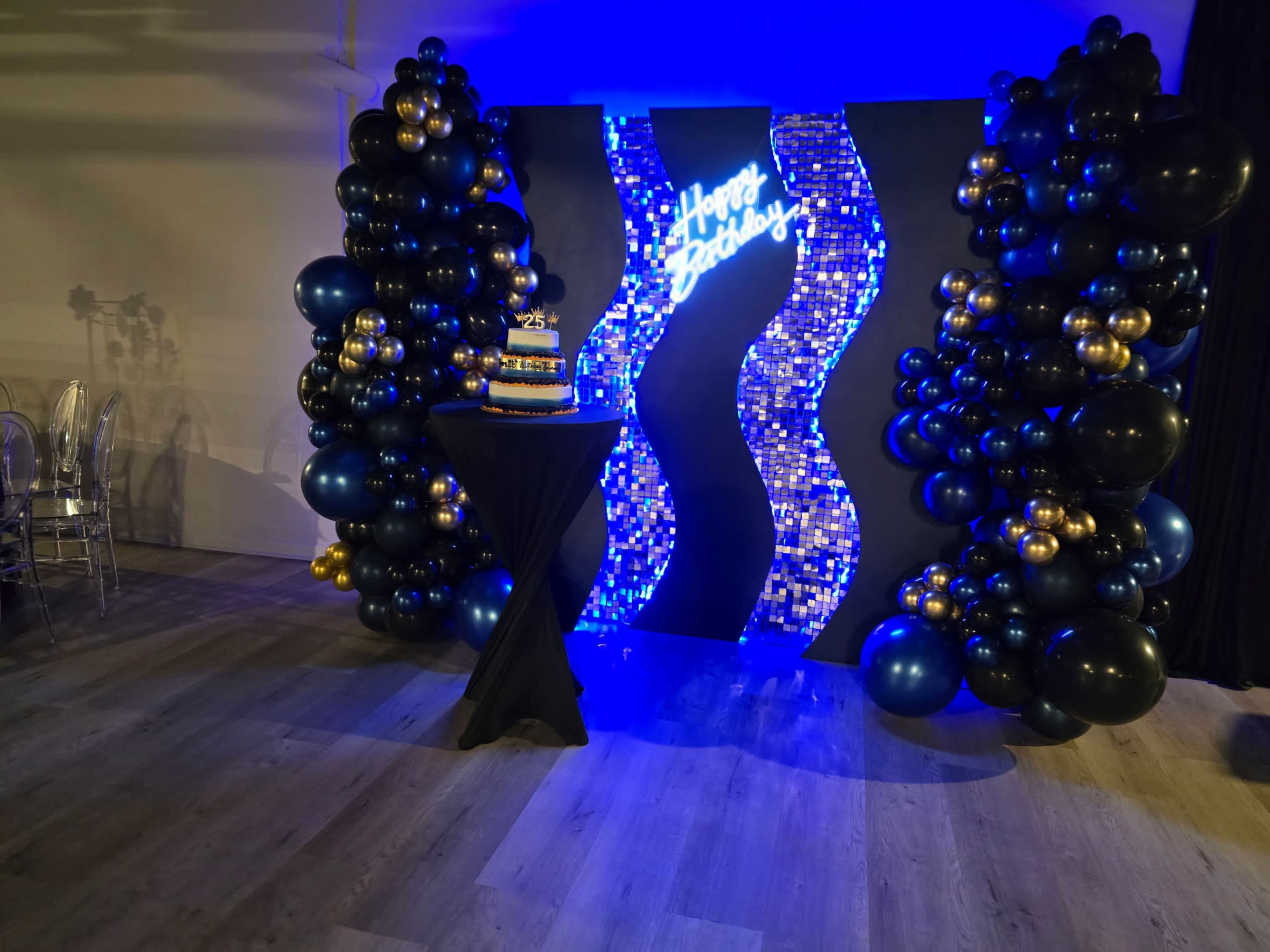 The image features a decorated event space with a balloon arch in black, gold, and blue around a stylish backdrop that displays "Happy Birthday," along with a birthday cake placed on a table.