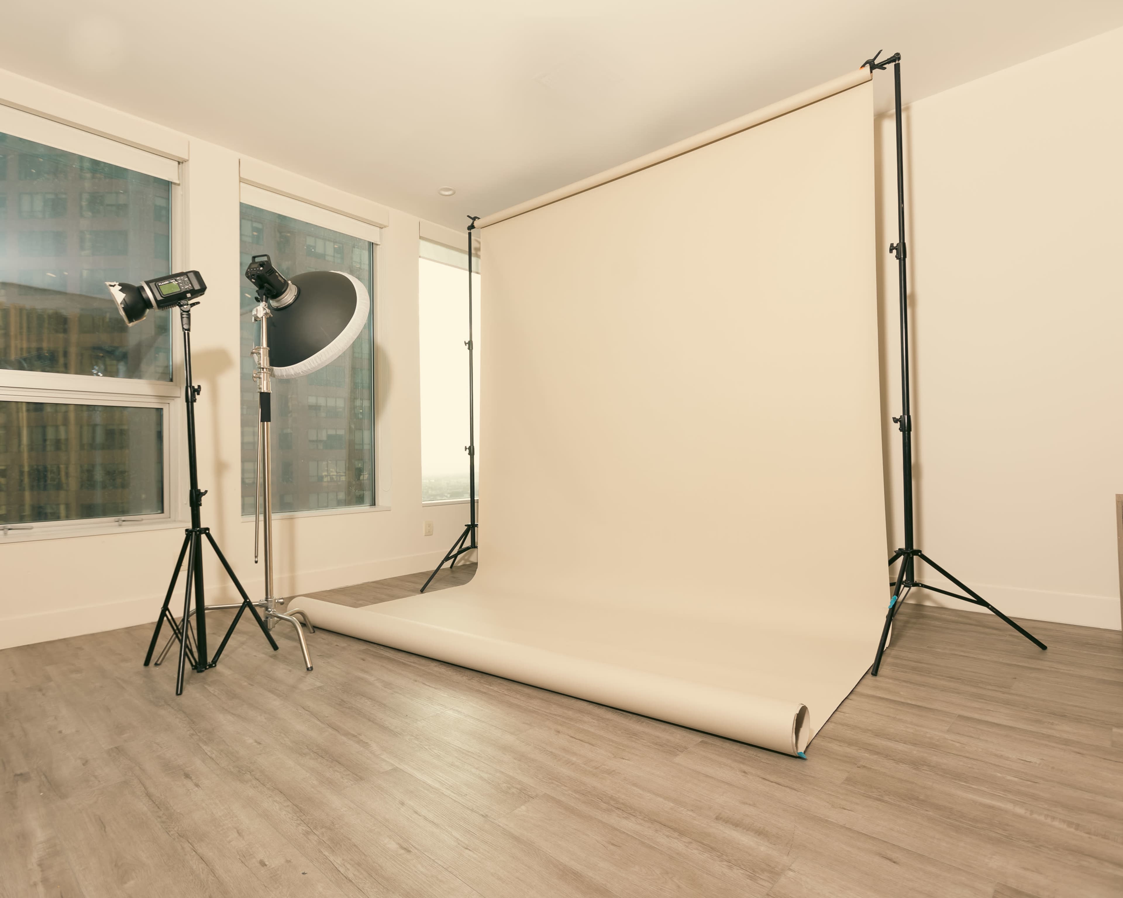 A photography studio with a beige backdrop set up on a smooth floor, accompanied by two studio lights and large windows in the background.