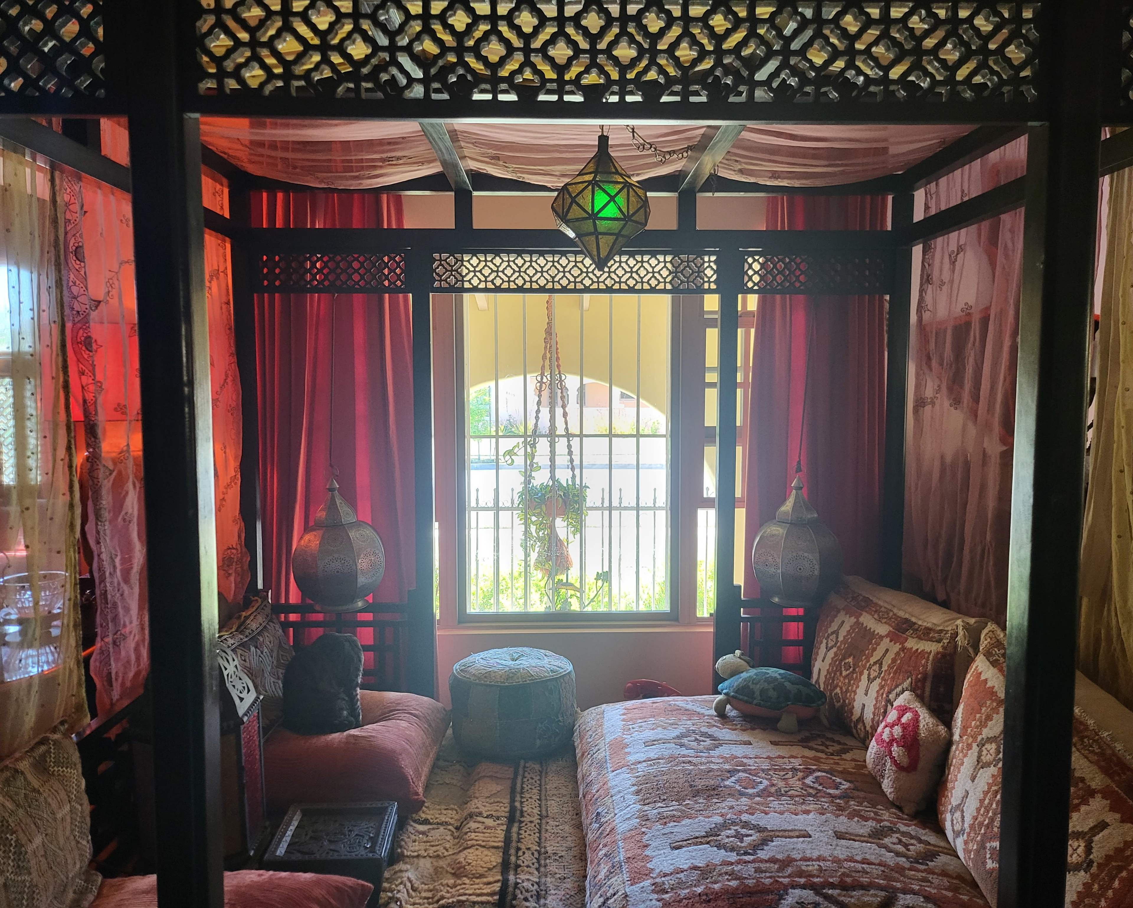 The image shows a cozy, elaborately decorated room featuring a four-poster bed with colorful textiles, lanterns, and patterned drapes, illuminated by natural light through a window.
