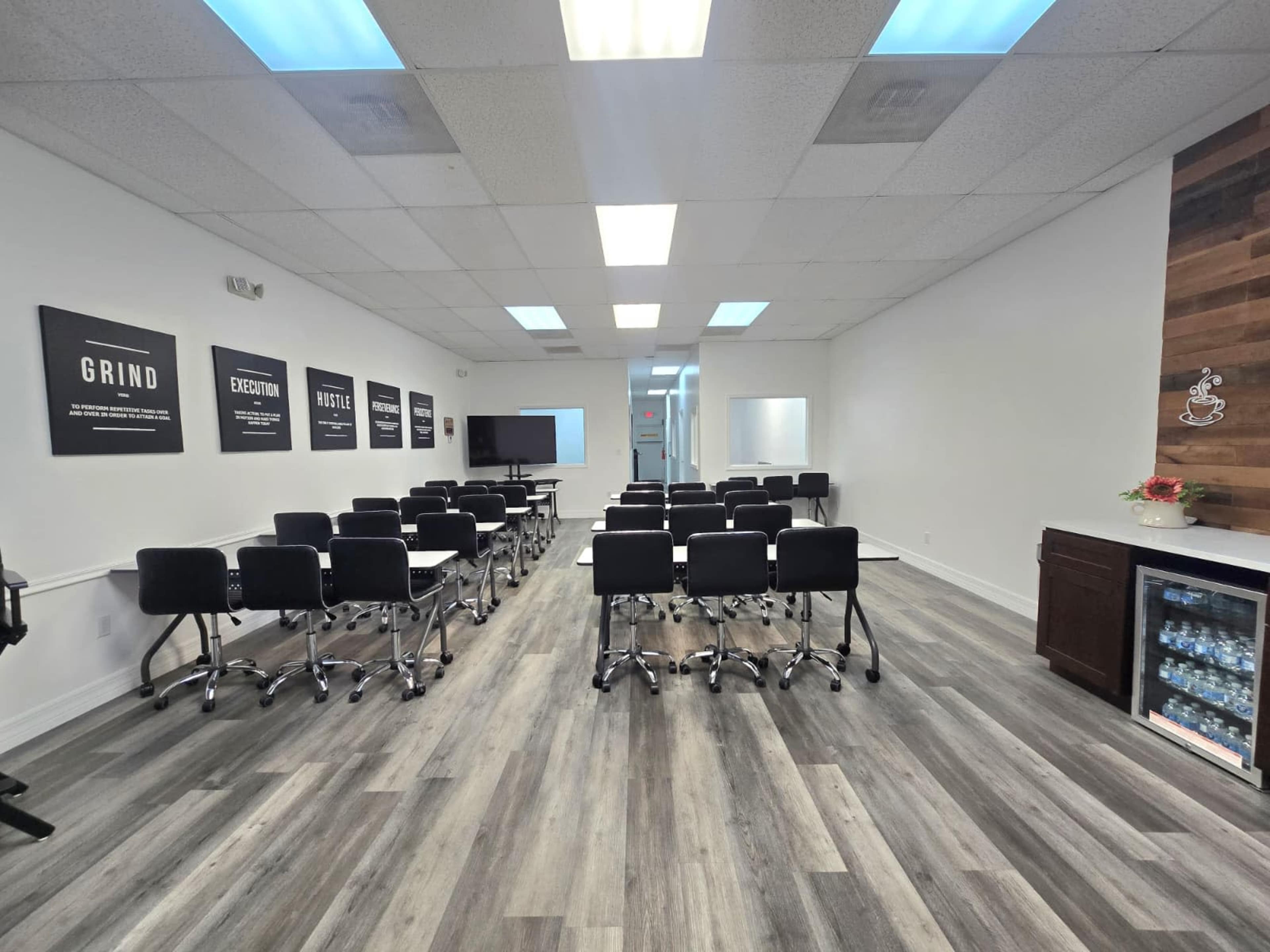 The image shows a large, modern training room set up with rows of black chairs and tables, along with motivational posters on the walls and a refreshment area on the side.