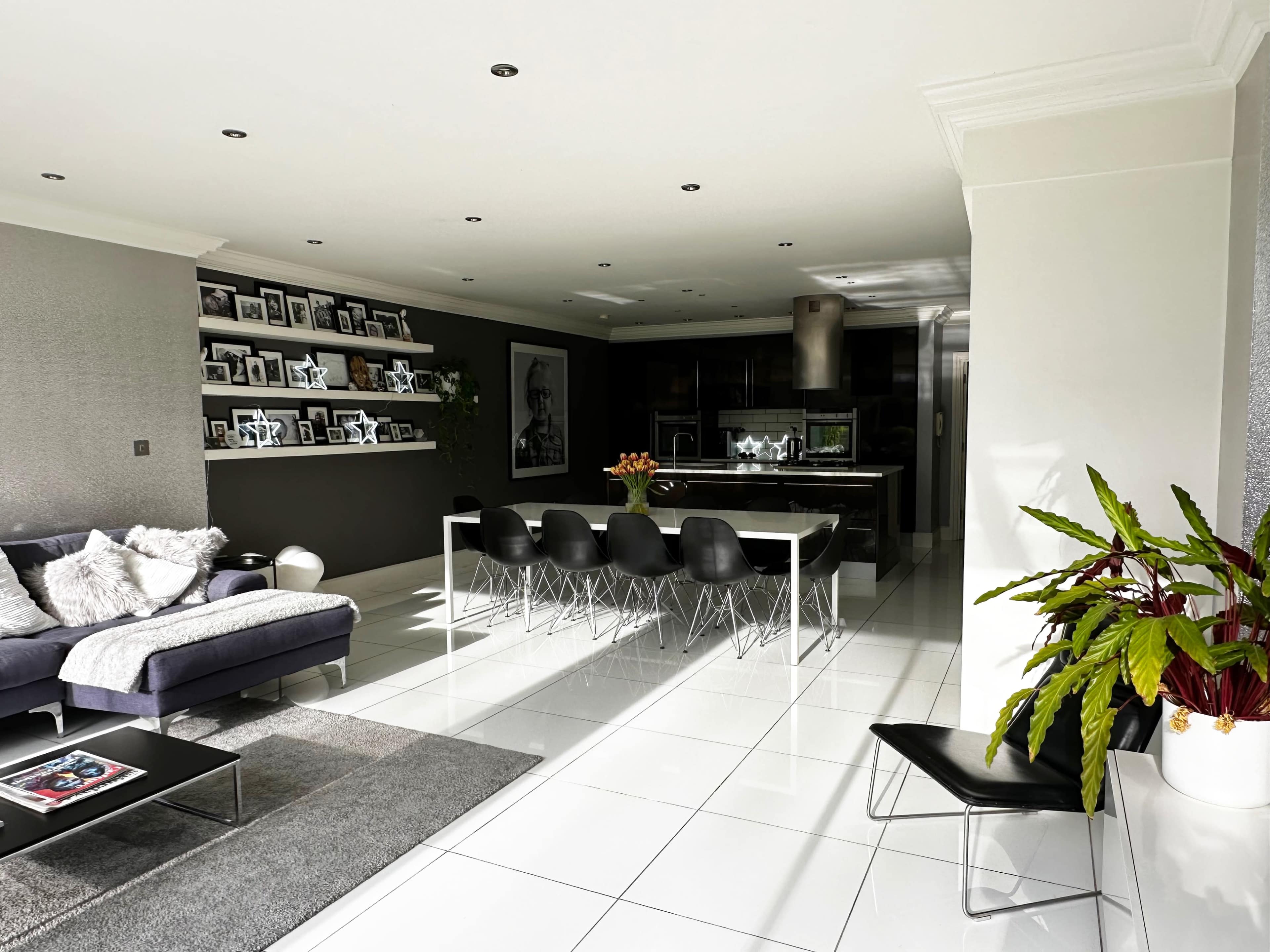 A modern living space features a grey sofa, a large dining table with black chairs, and shelves adorned with framed photographs.