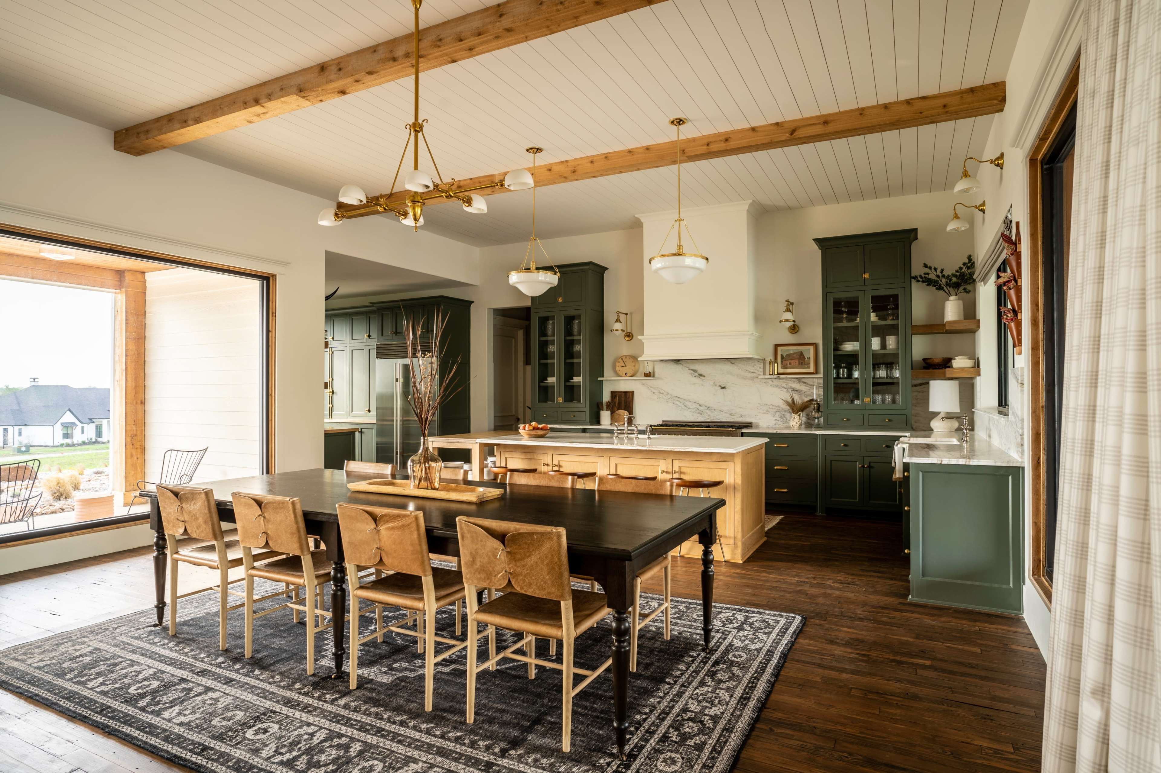 A spacious kitchen features a wooden dining table surrounded by chairs, with green cabinetry, marble countertops, and large windows overlooking a patio.