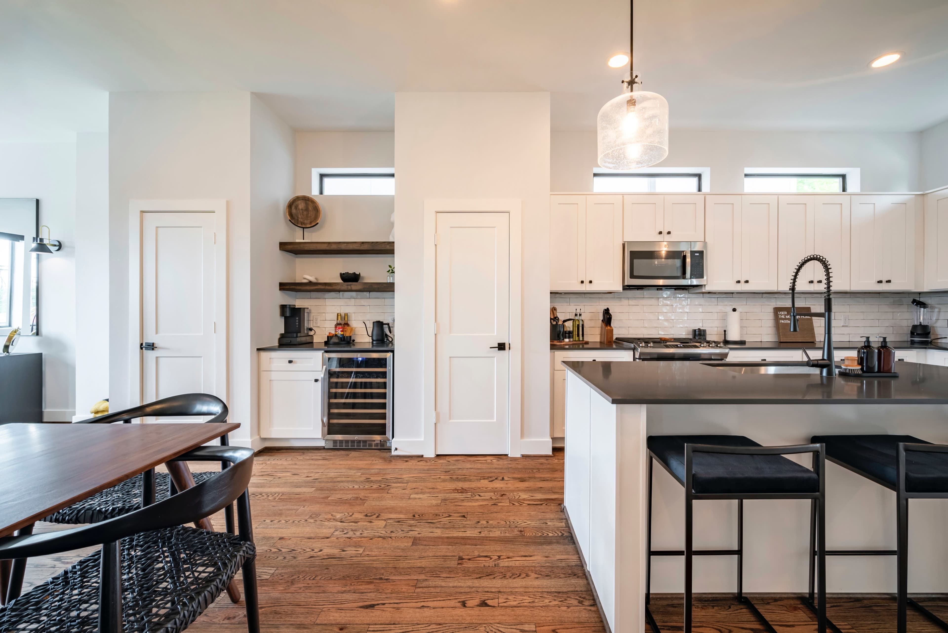 A modern kitchen with white cabinets, stainless steel appliances, a dark countertop, and wooden flooring, featuring a dining table nearby.