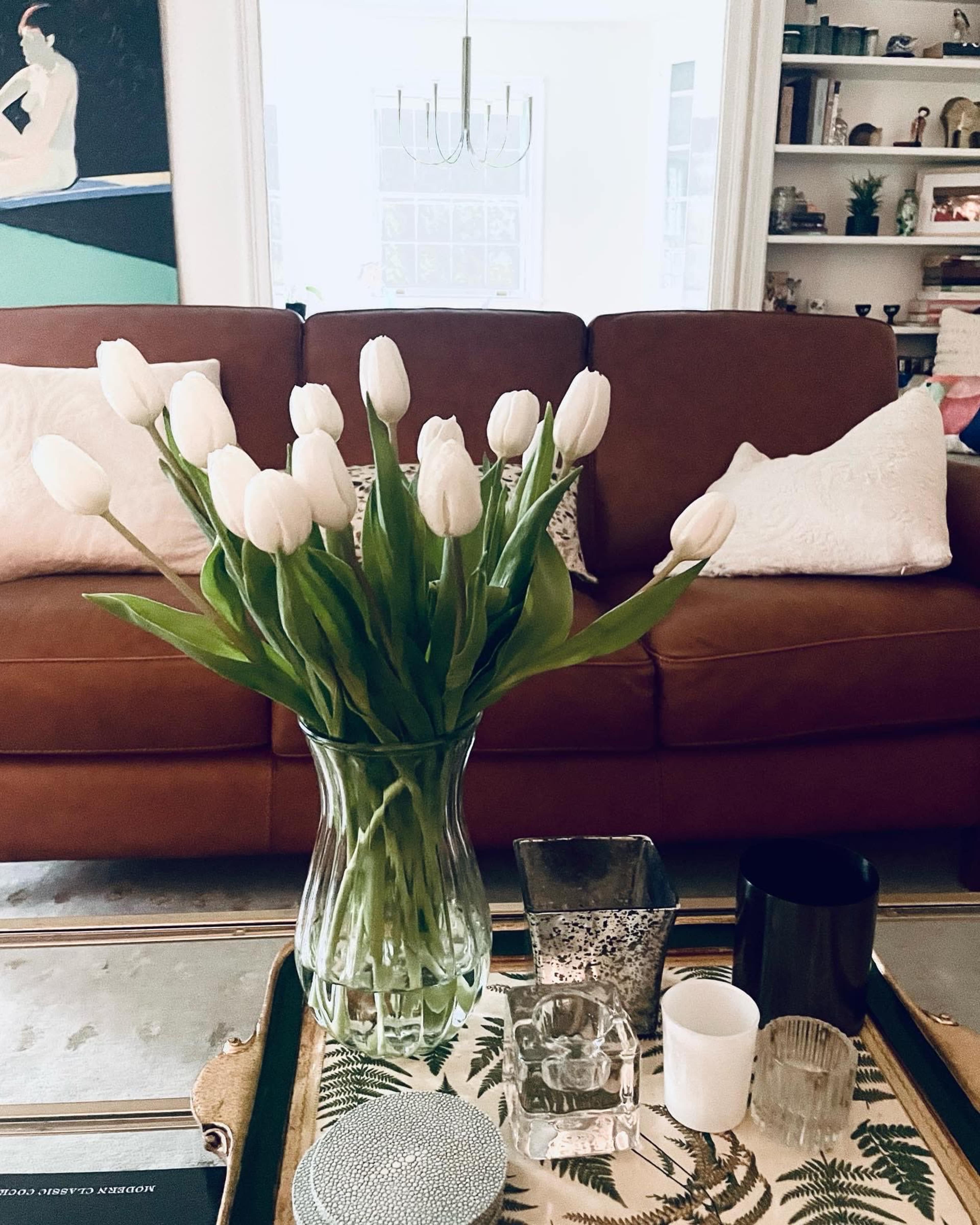 A vase of white tulips sits on a coffee table in a living room with a brown couch and decorative items.