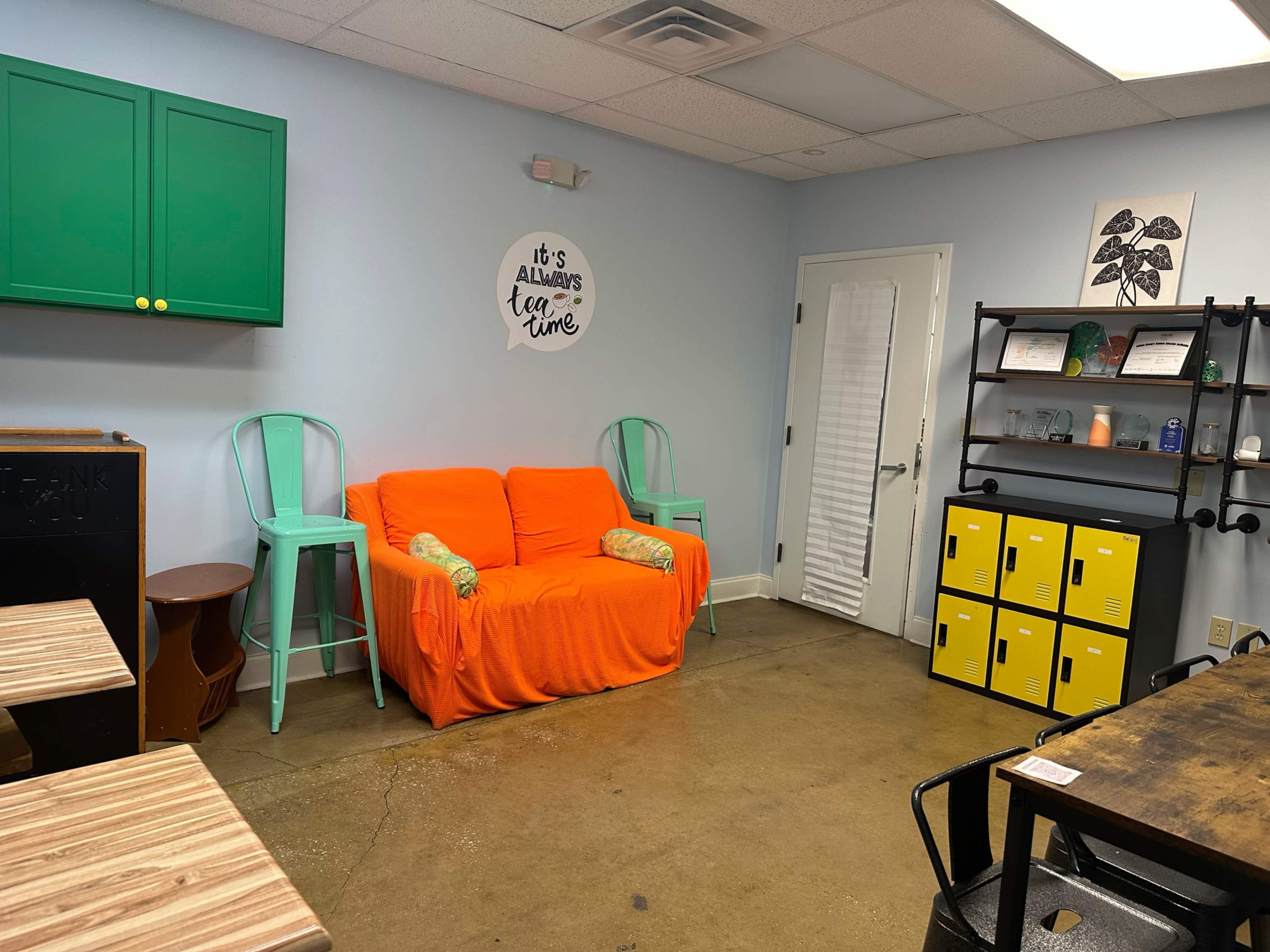 The image shows a small, brightly colored room featuring an orange couch, two teal chairs, a black shelf with yellow lockers, and a wall with a decorative sign.