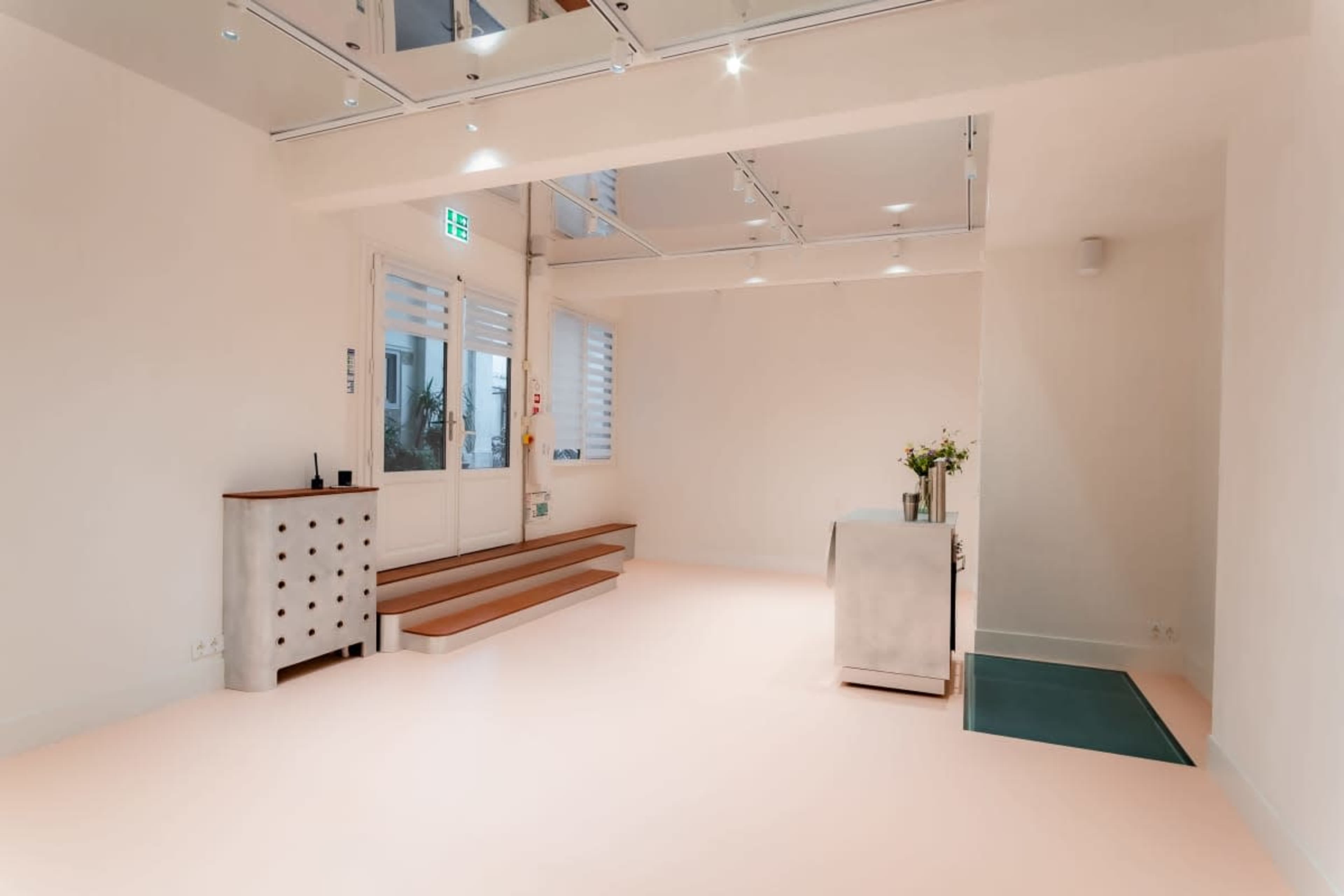 The image shows a minimalist interior space with light-colored walls, a flat floor, a reception desk, and a small staircase.