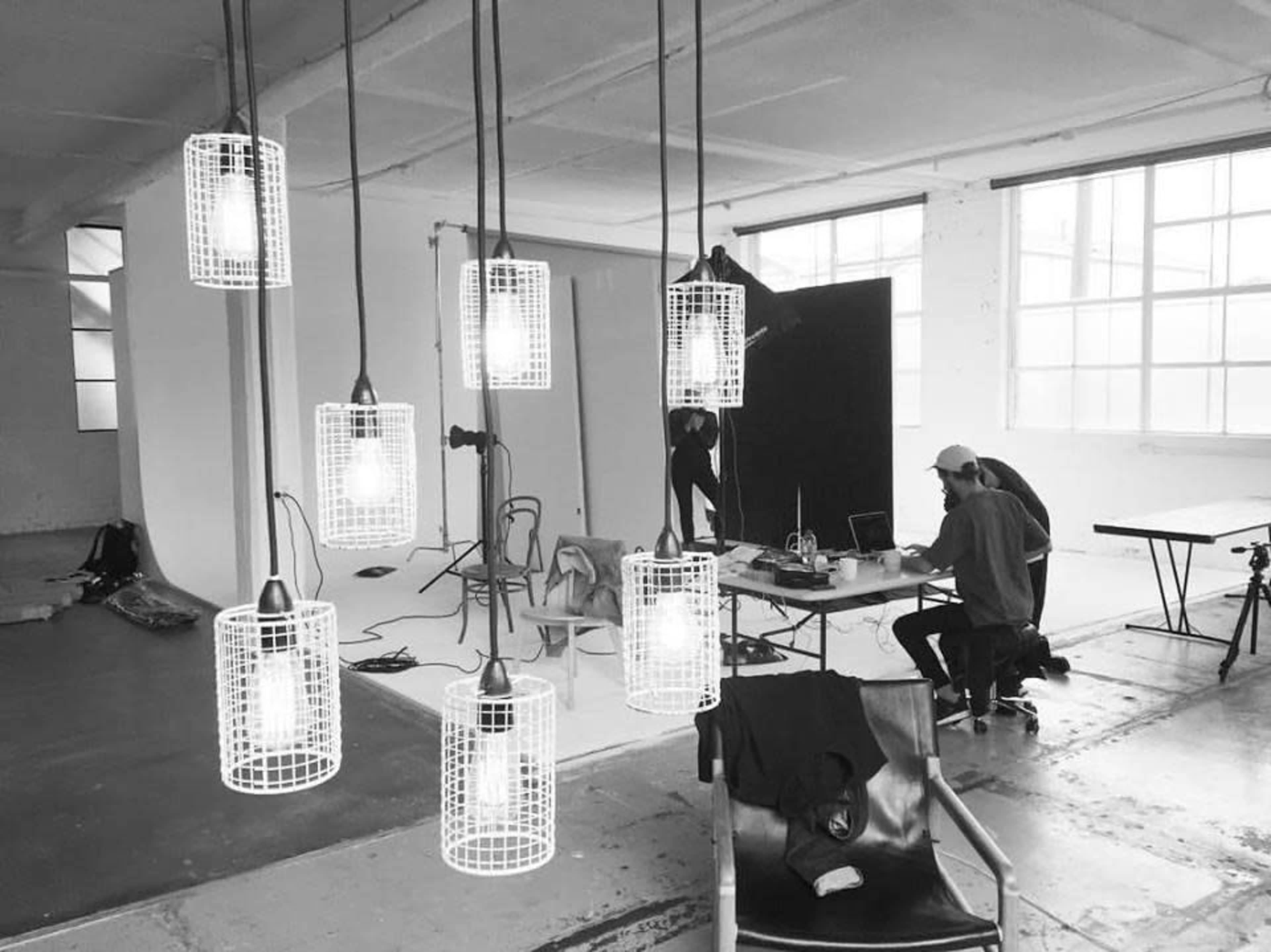 A spacious studio is illuminated by hanging pendant lights as two people work at a table while a backdrop is set up nearby.