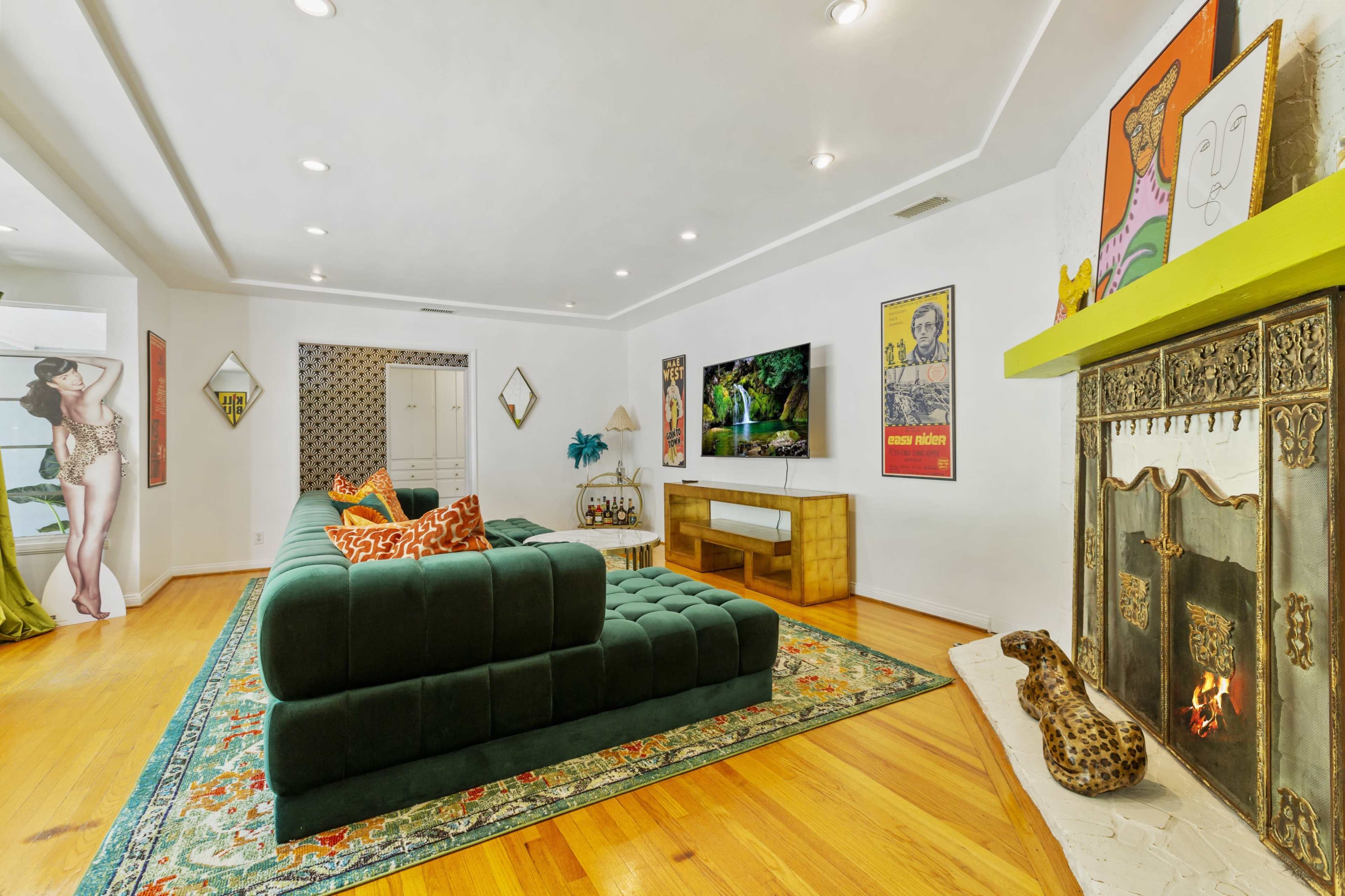 The living room features a green sectional sofa, a large television mounted on a wall, a decorative fireplace with a leopard figurine, and various art pieces displayed throughout the space.