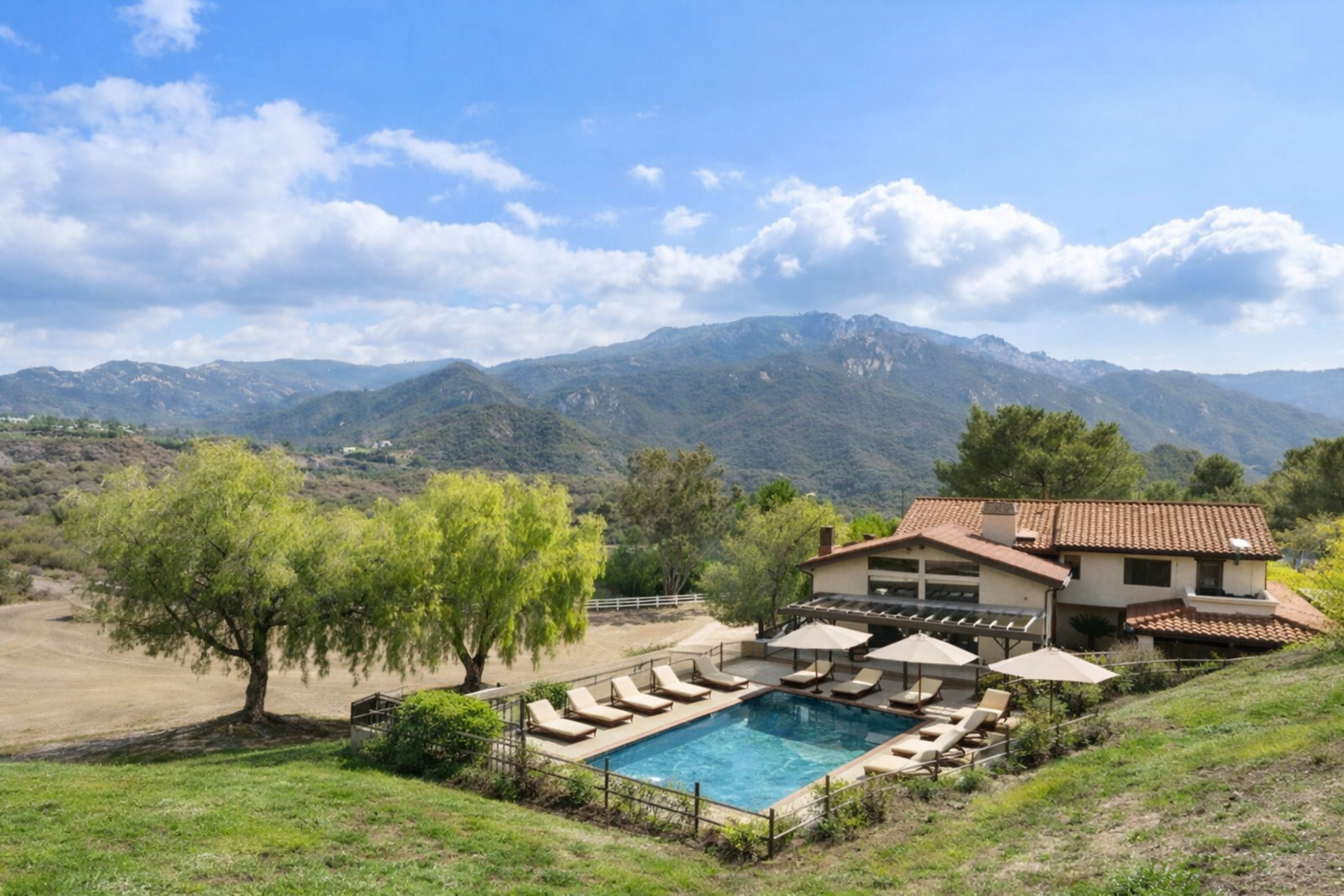 A modern villa with a swimming pool and lounge chairs is set against a backdrop of mountains and blue skies.
