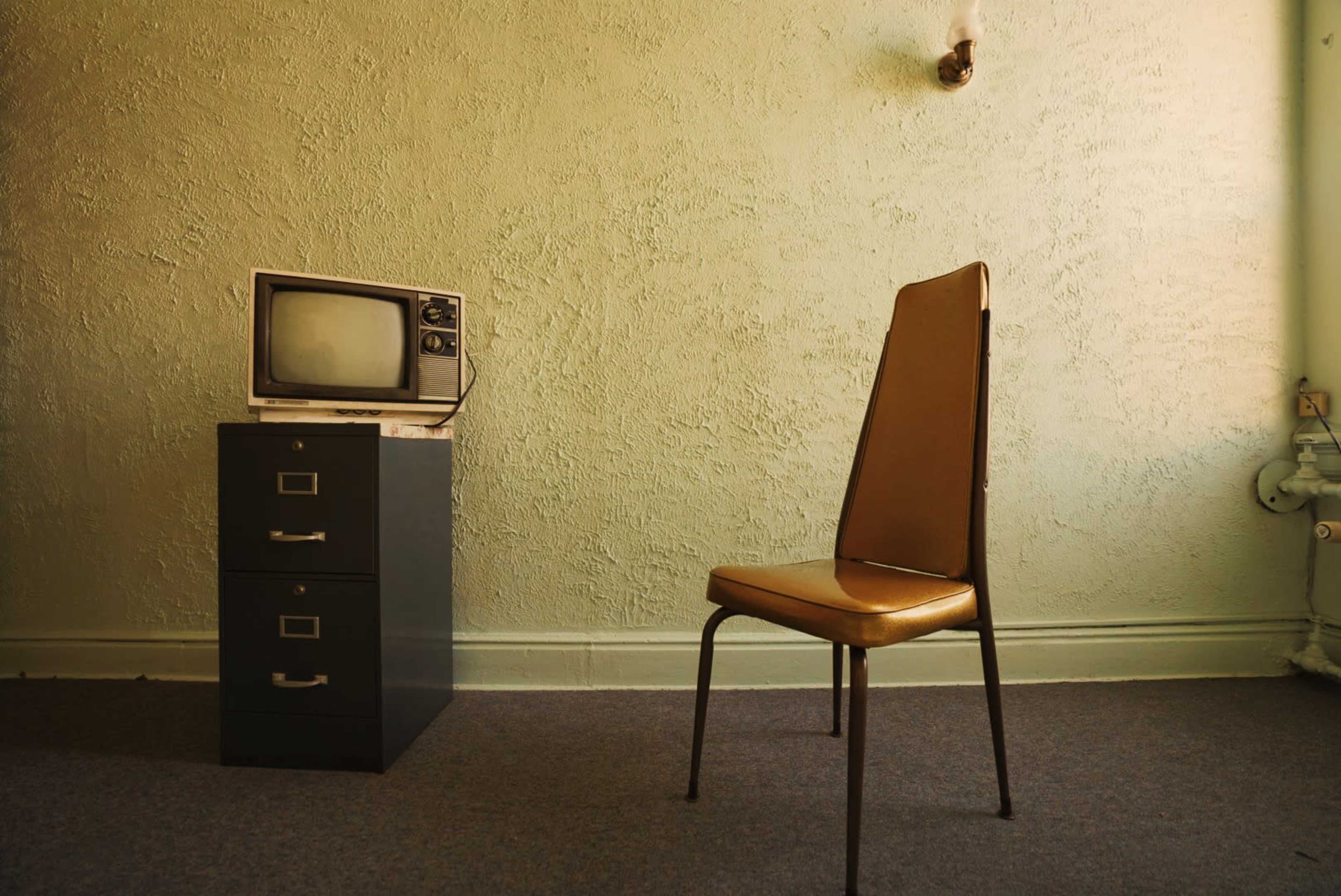 A small room with a green textured wall, a vintage television on top of a black filing cabinet, and a single brown chair positioned nearby.