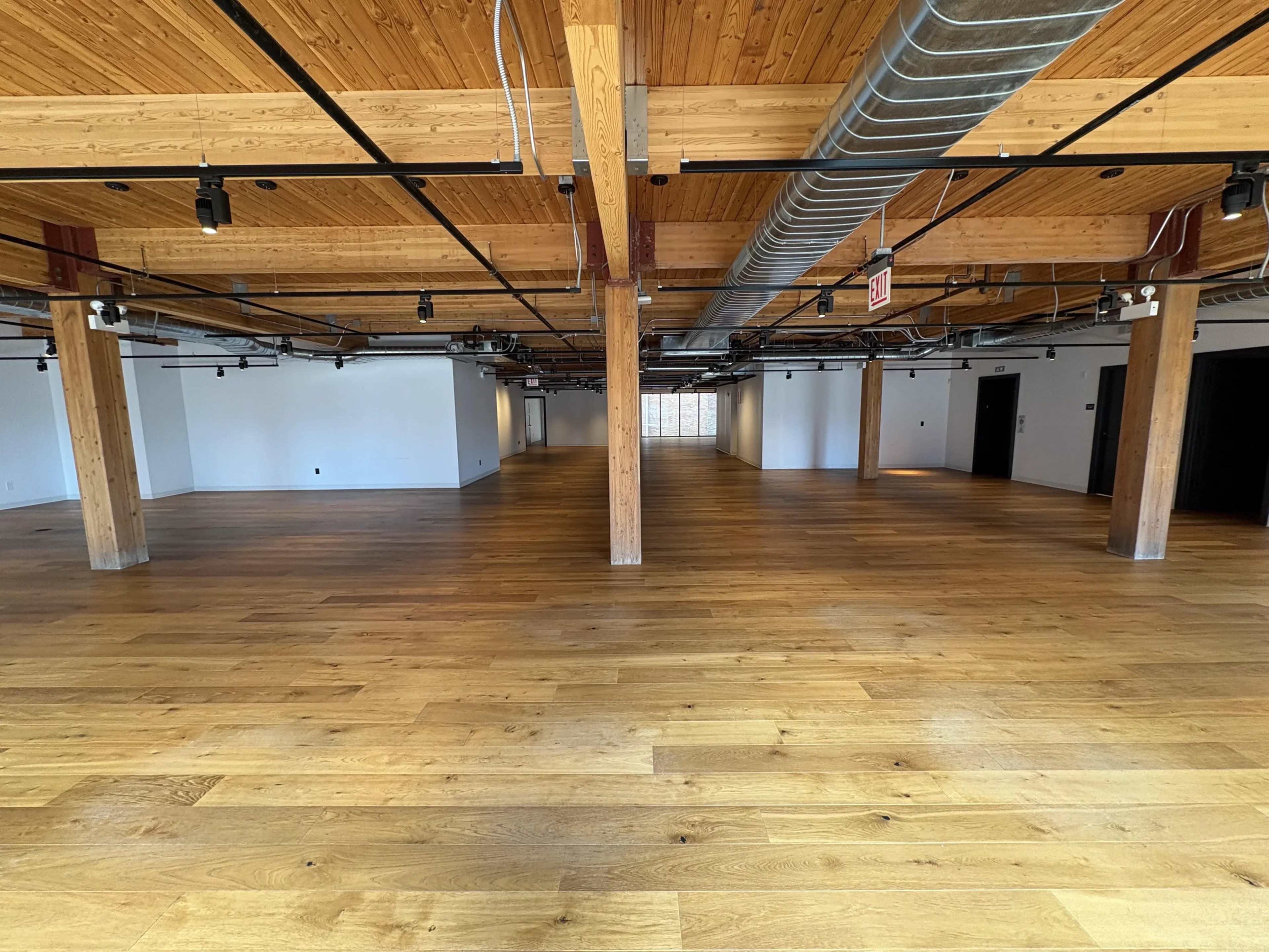 The image shows a spacious, empty interior with wooden beams and flooring, featuring exposed pipes and track lighting.