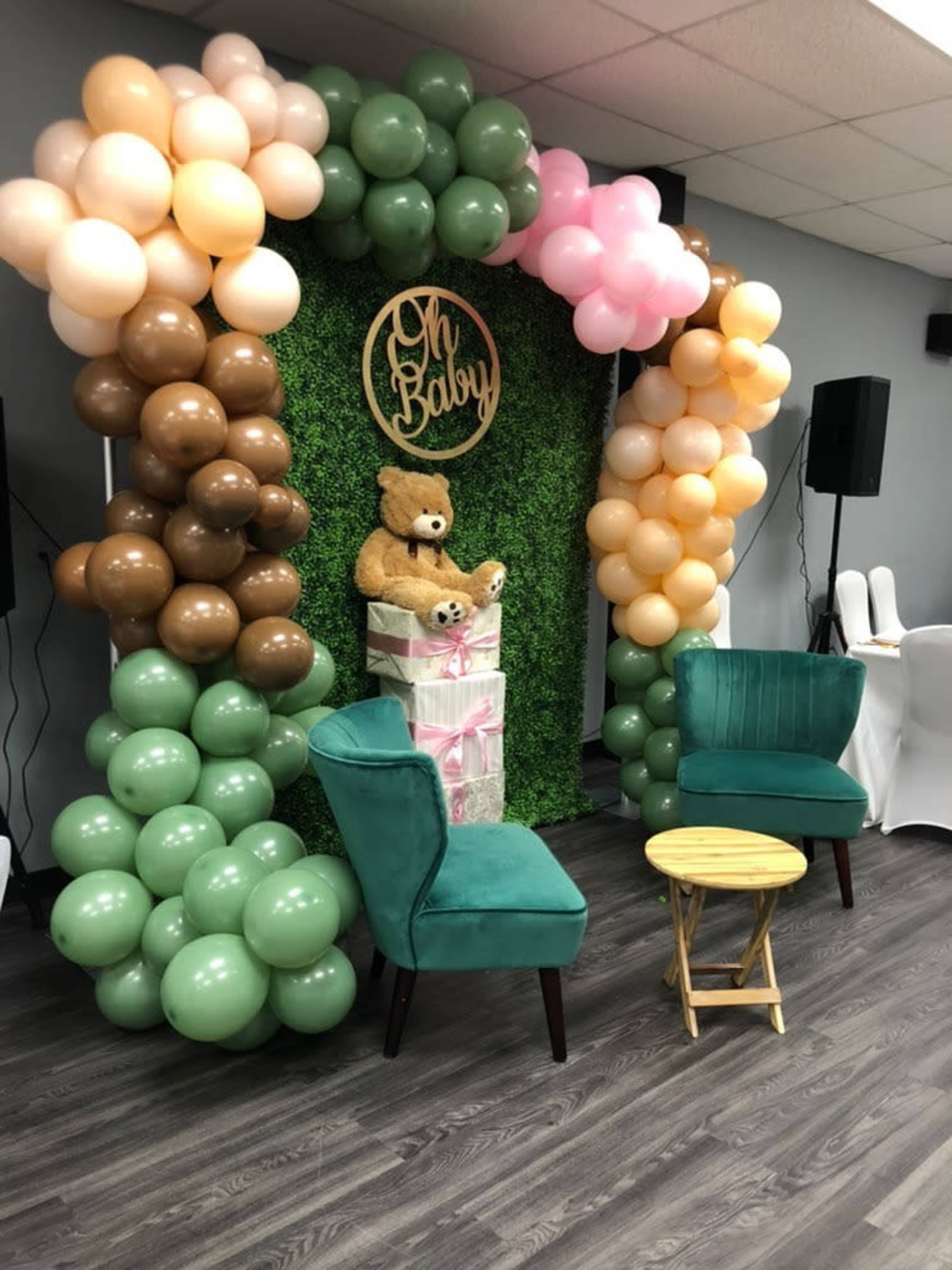 A baby shower backdrop displays a greenery wall adorned with a teddy bear and a "Oh Baby" sign, framed by a balloon arrangement in pastel colors and accompanied by two green chairs and a small wooden table.