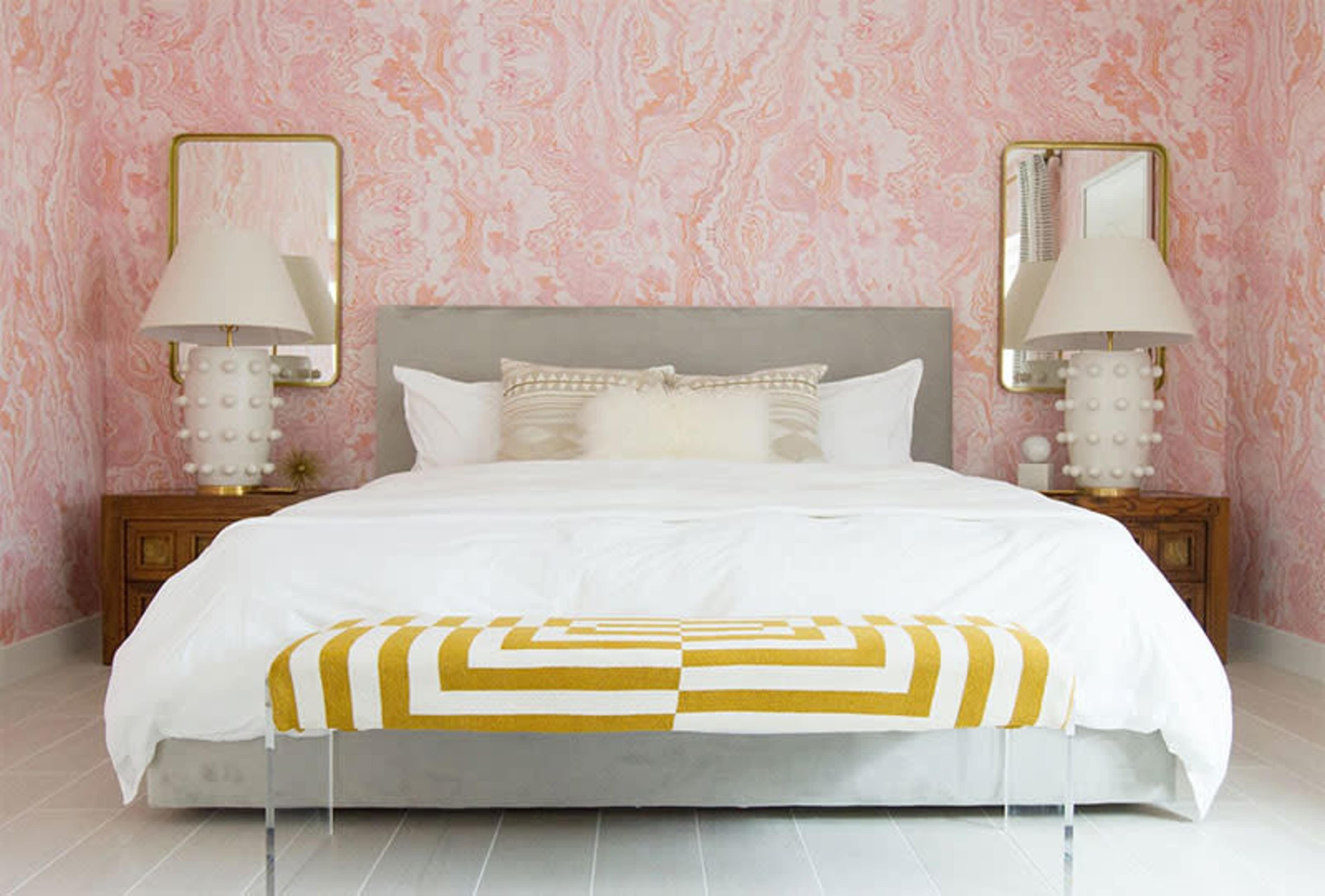 A neatly arranged bedroom featuring a large bed with a white comforter, flanked by two lamps and mirrors against a pink marbled wallpaper.