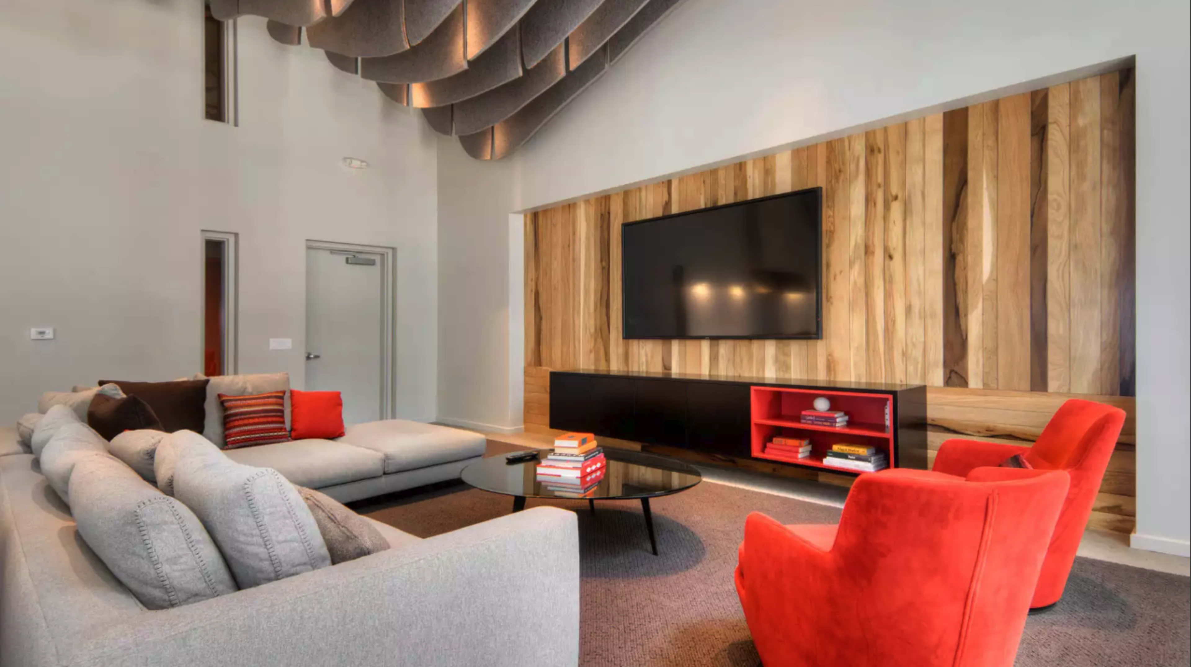 A modern living room features a sectional sofa, two red chairs, a glass coffee table, and a large wall-mounted television above a wooden accent wall.