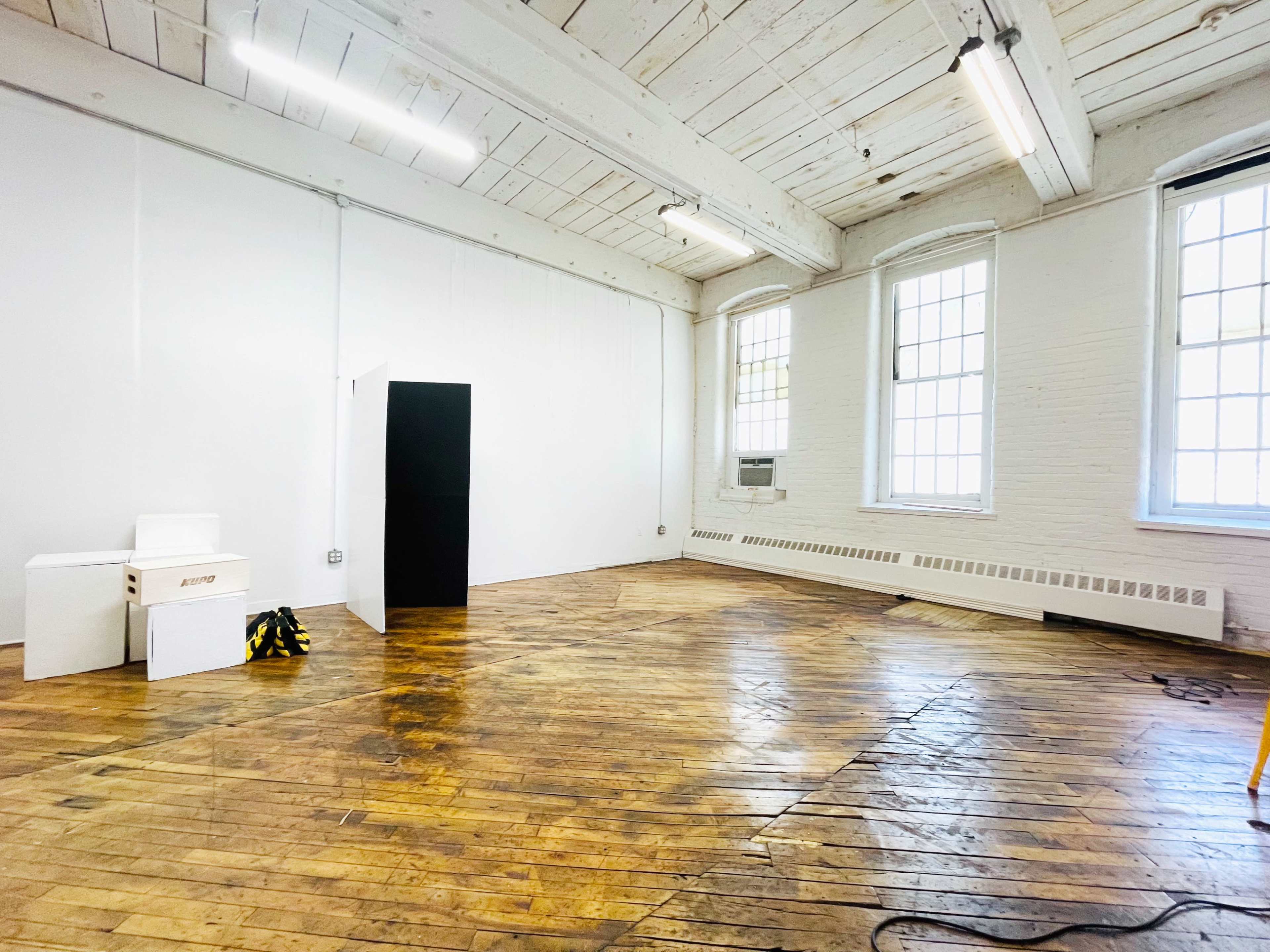 An empty, well-lit room with wooden floors, large windows, and minimal furnishings, including a black storage unit and white boxes.