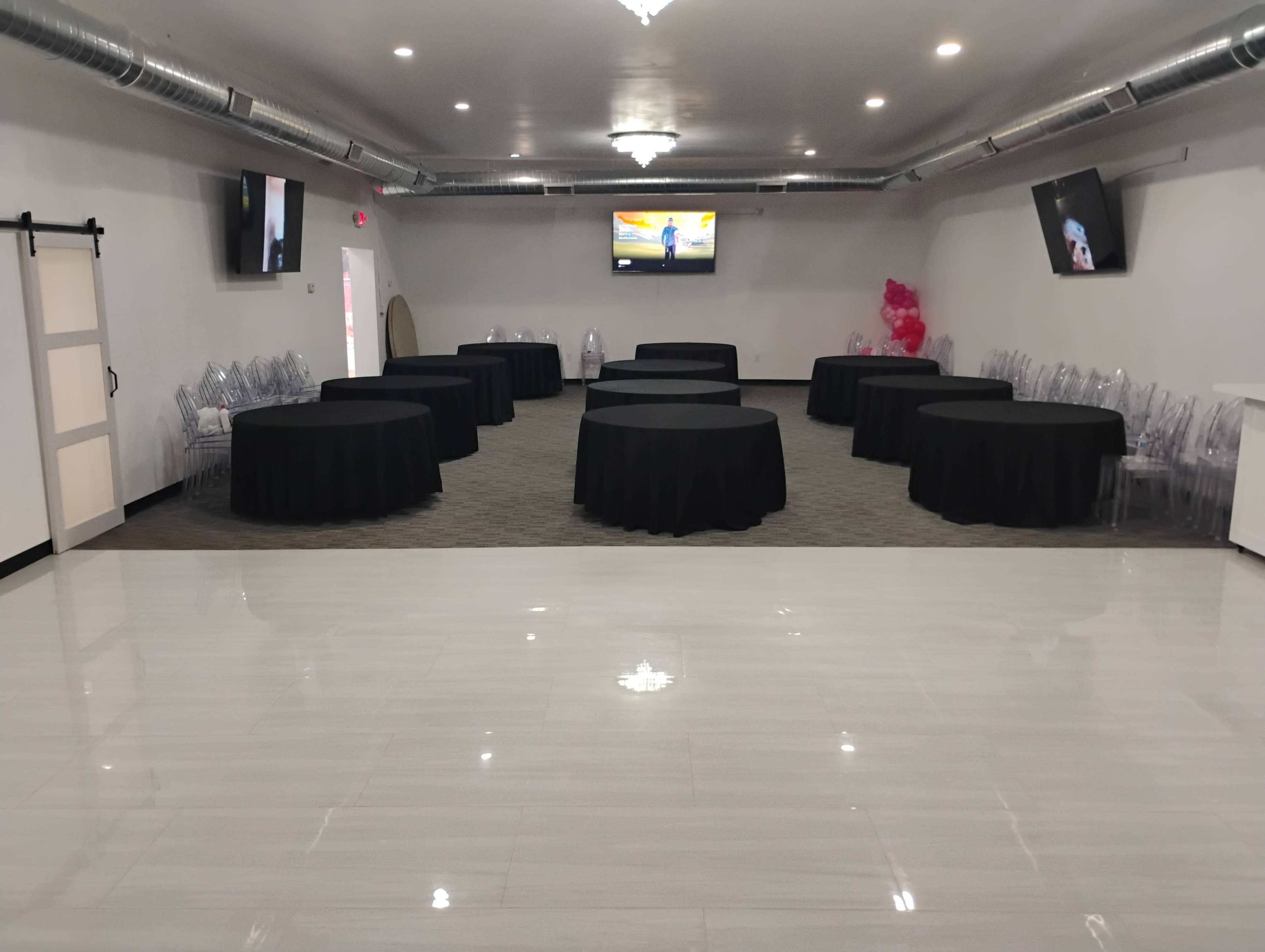 The image shows a spacious event venue with multiple round tables covered in black tablecloths and two televisions mounted on the walls.