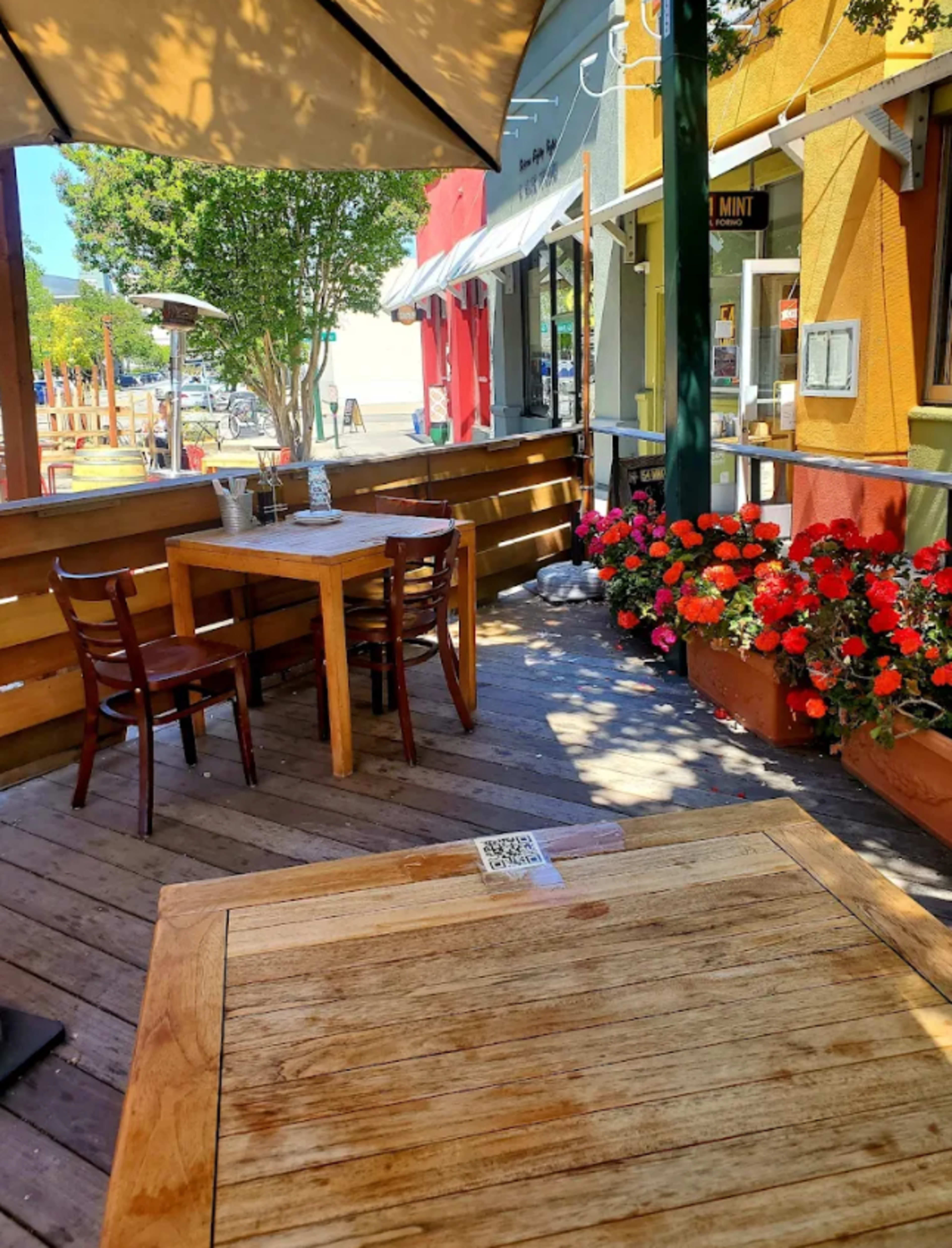 A small outdoor dining area features wooden tables and chairs surrounded by colorful flowers and shaded by an umbrella.