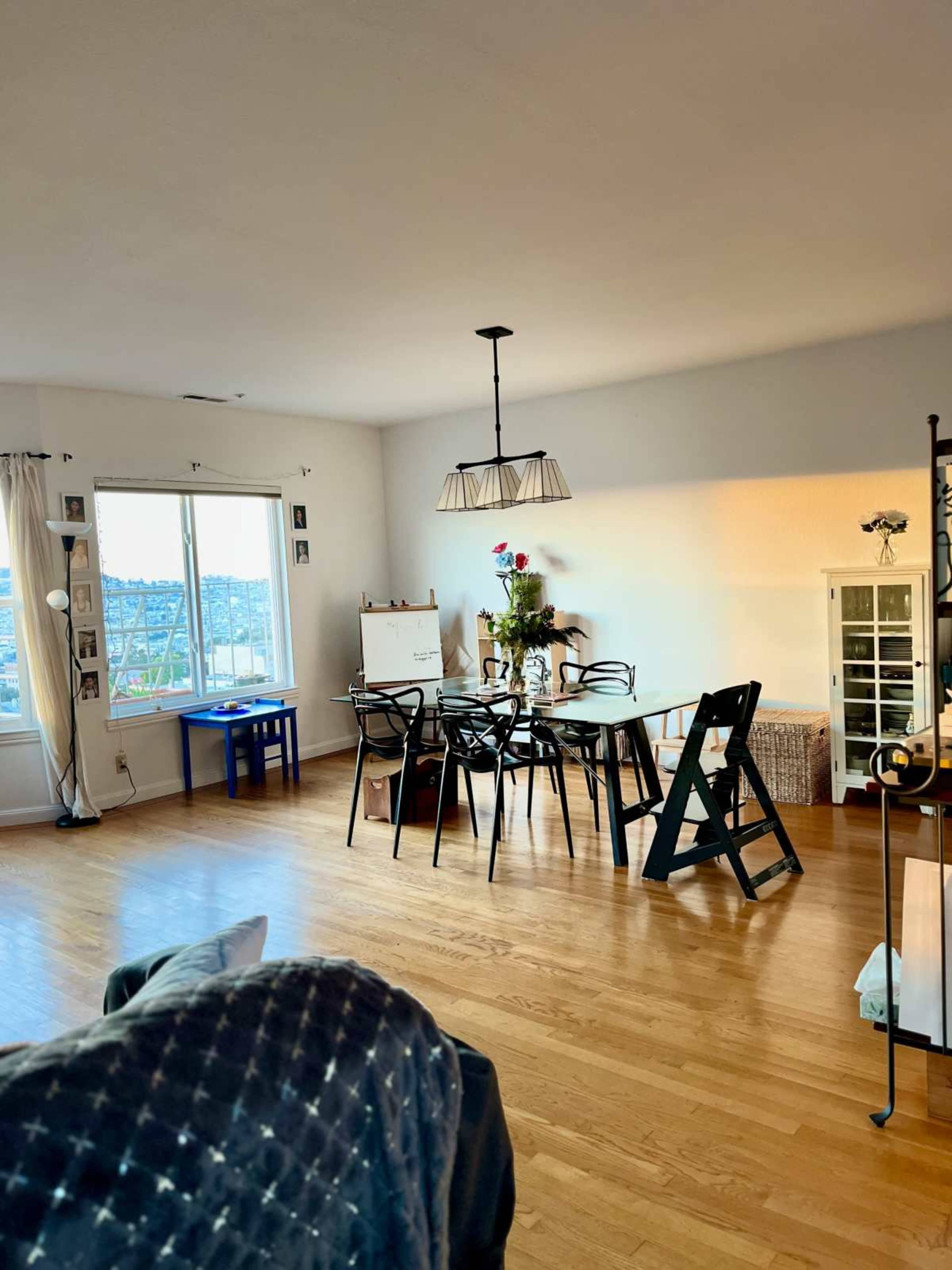 The image shows a brightly lit dining area with a wooden floor, a table surrounded by black chairs, a window with a view, and various decorative items on shelves.