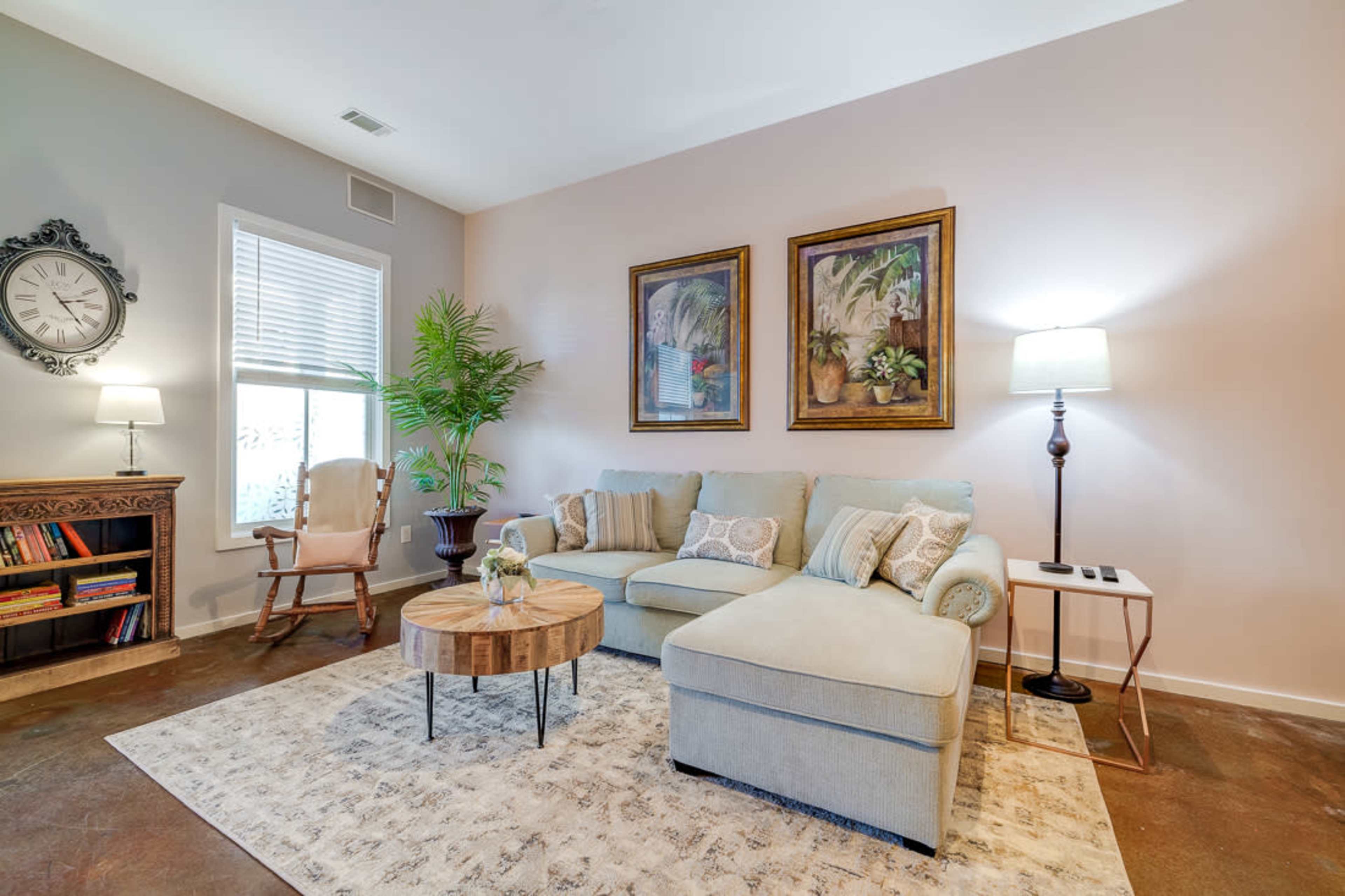 The image shows a living room featuring a light blue sectional sofa, two framed paintings on the wall, a round coffee table, and a rocking chair next to a window.