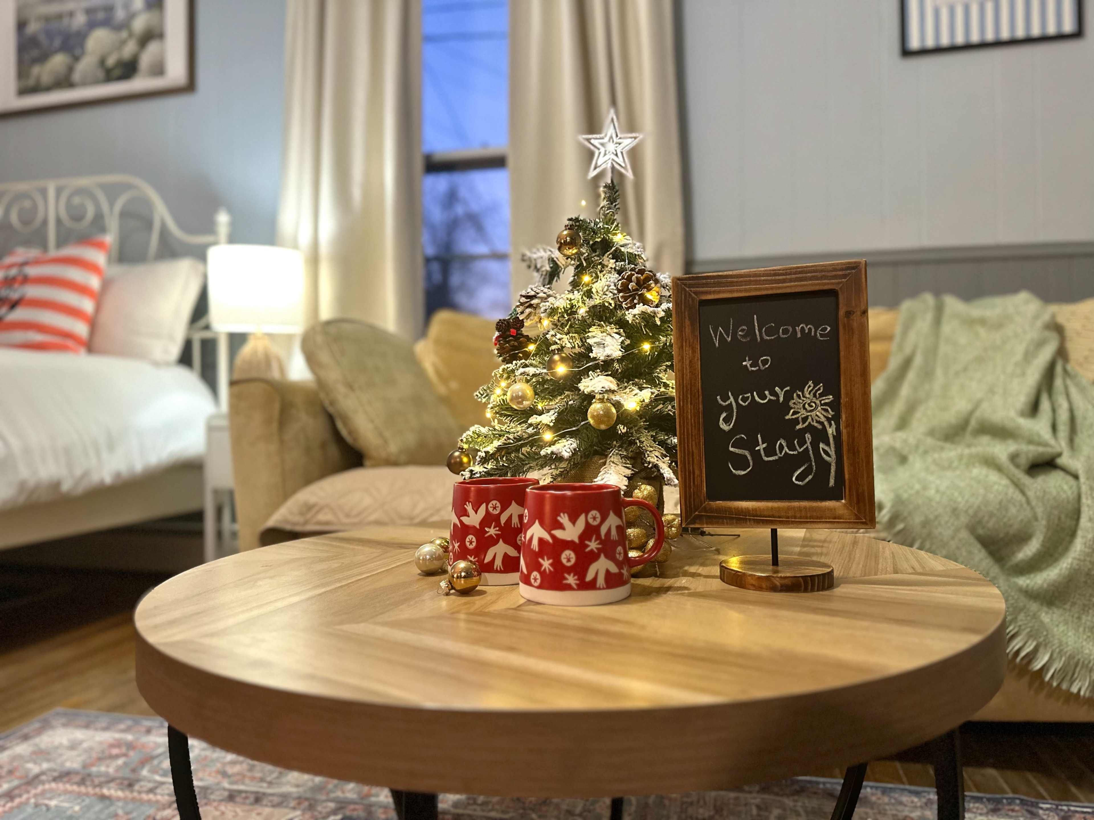 The scene features a cozy living room with a small decorated Christmas tree, a wooden coffee table with two red mugs, and a chalkboard sign that reads "Welcome to your Stay."