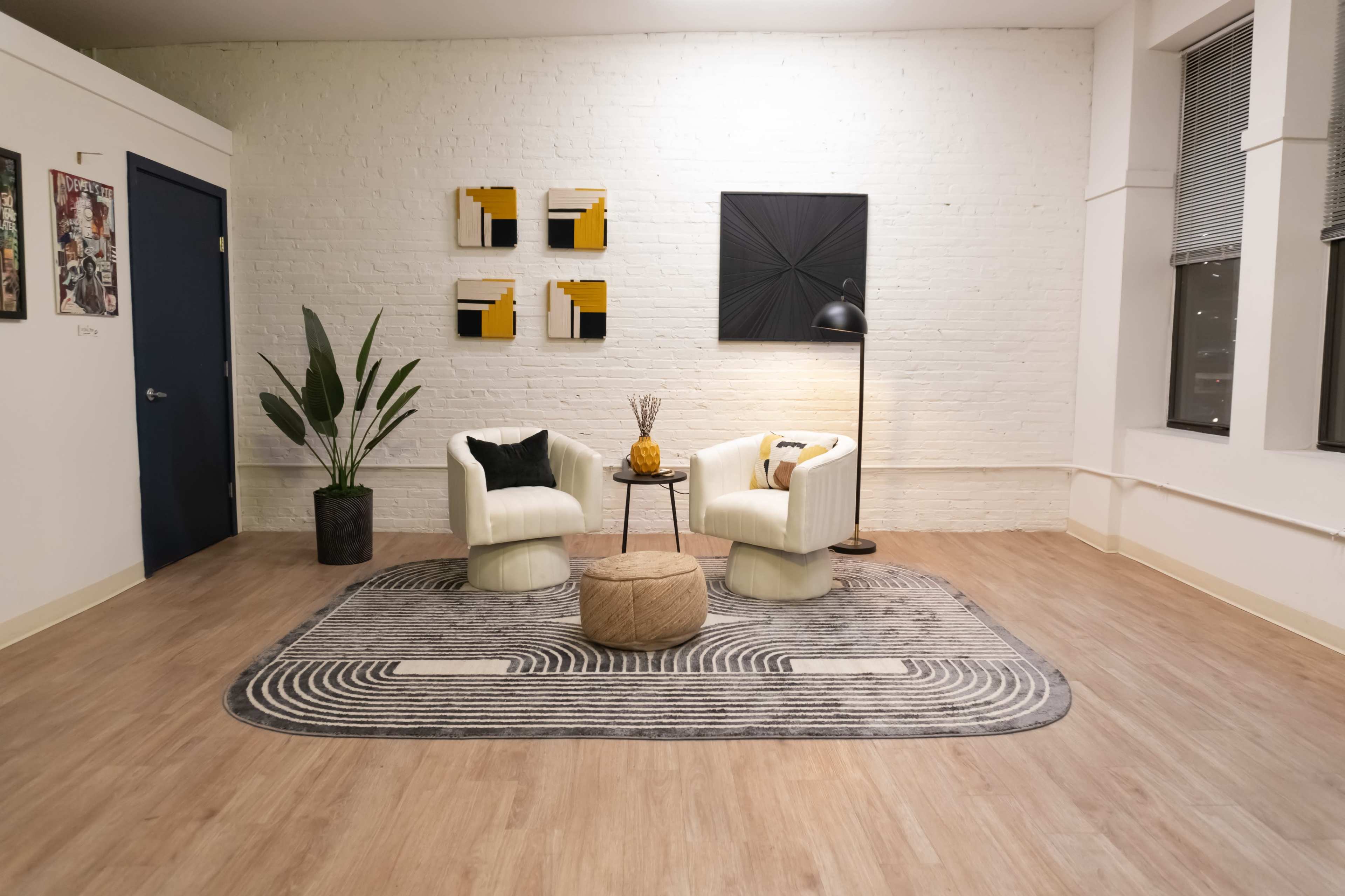 The image shows a minimalistic living space with two white upholstered chairs facing each other, a round ottoman, a tall plant in a black pot, and abstract wall art.