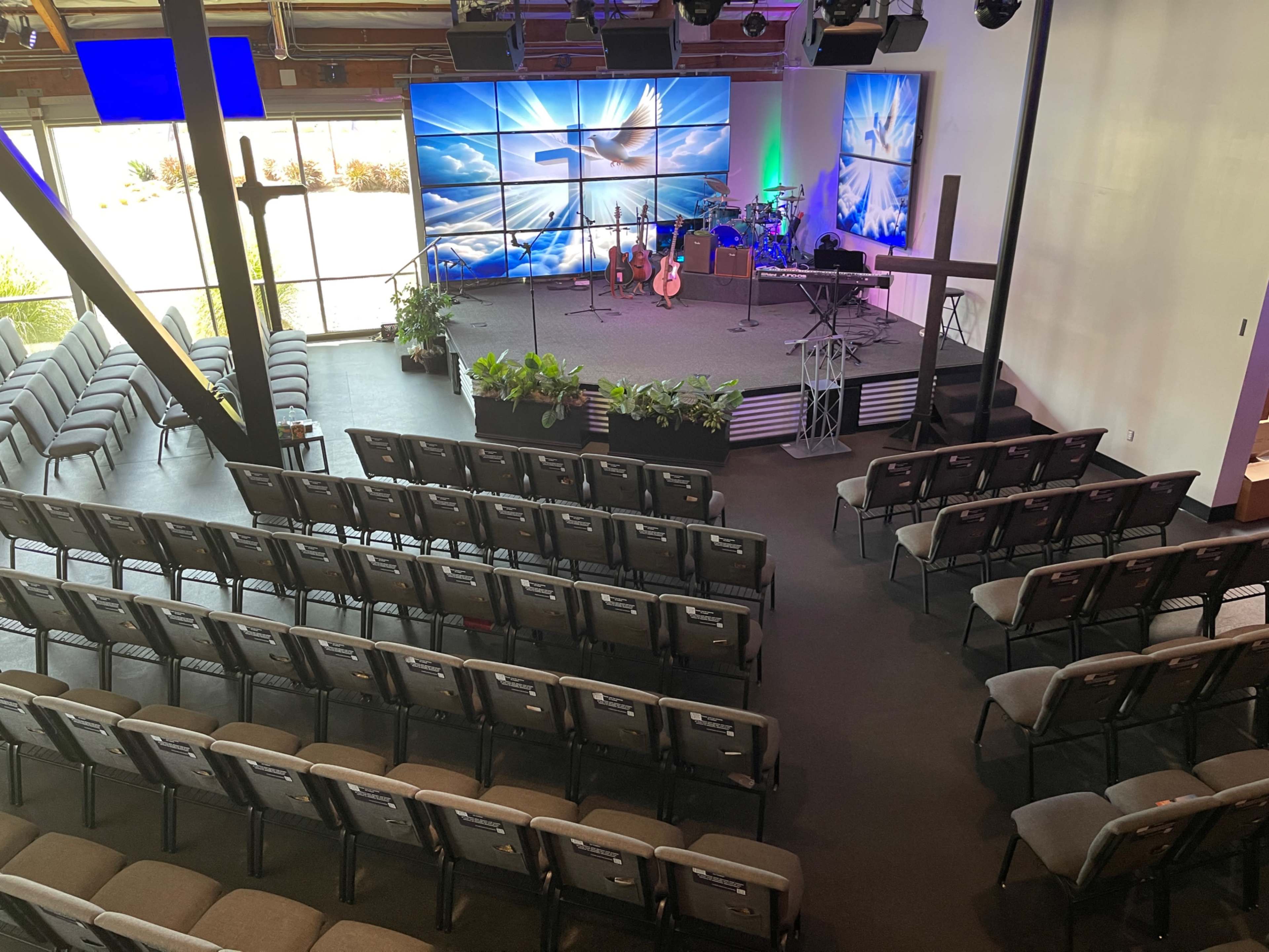 A spacious auditorium features rows of seating facing a stage with musical instruments and a cross, illuminated by large screens displaying clouds and light.