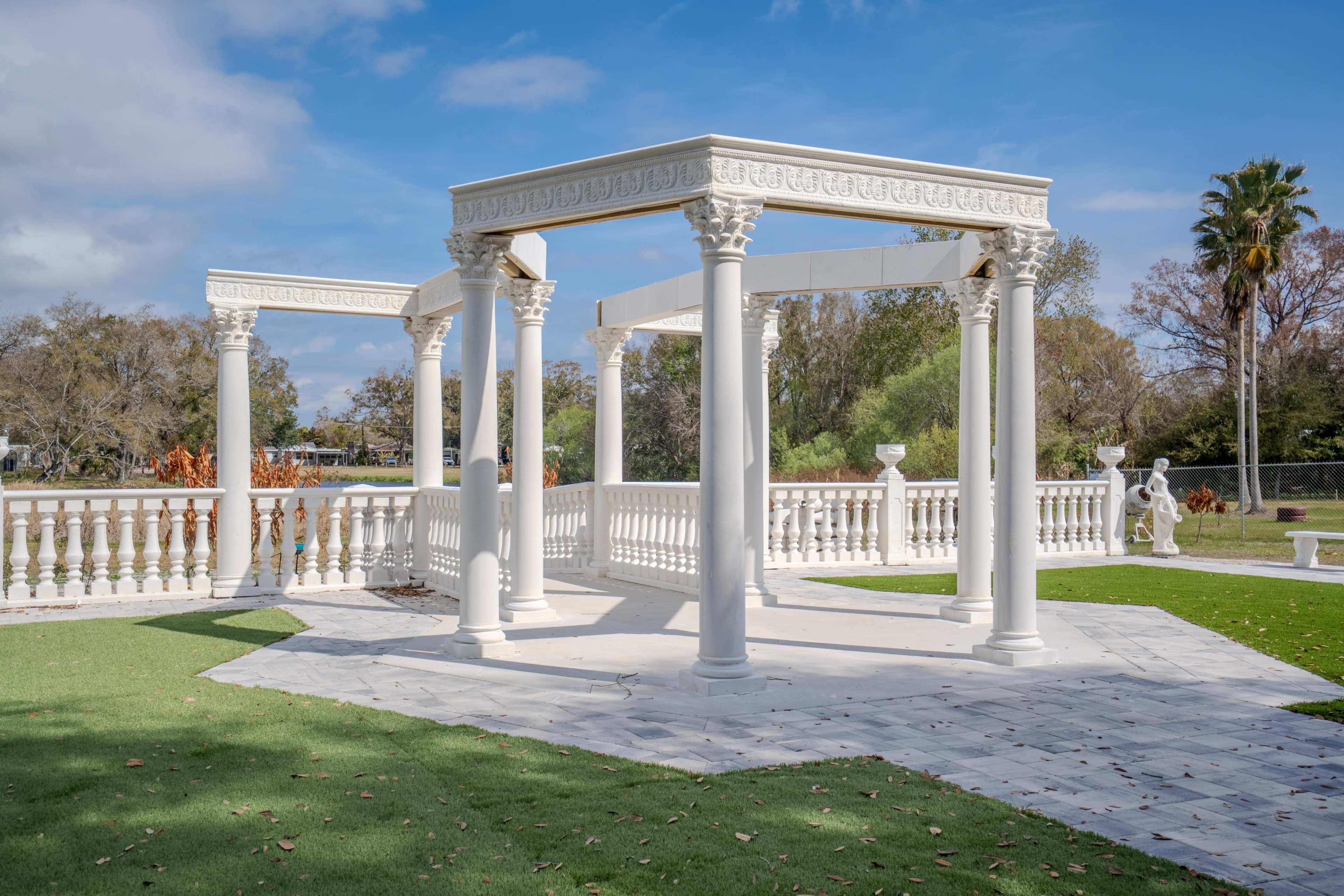 The image shows a white architectural structure with columns and an open canopy situated in a landscaped area with green grass.