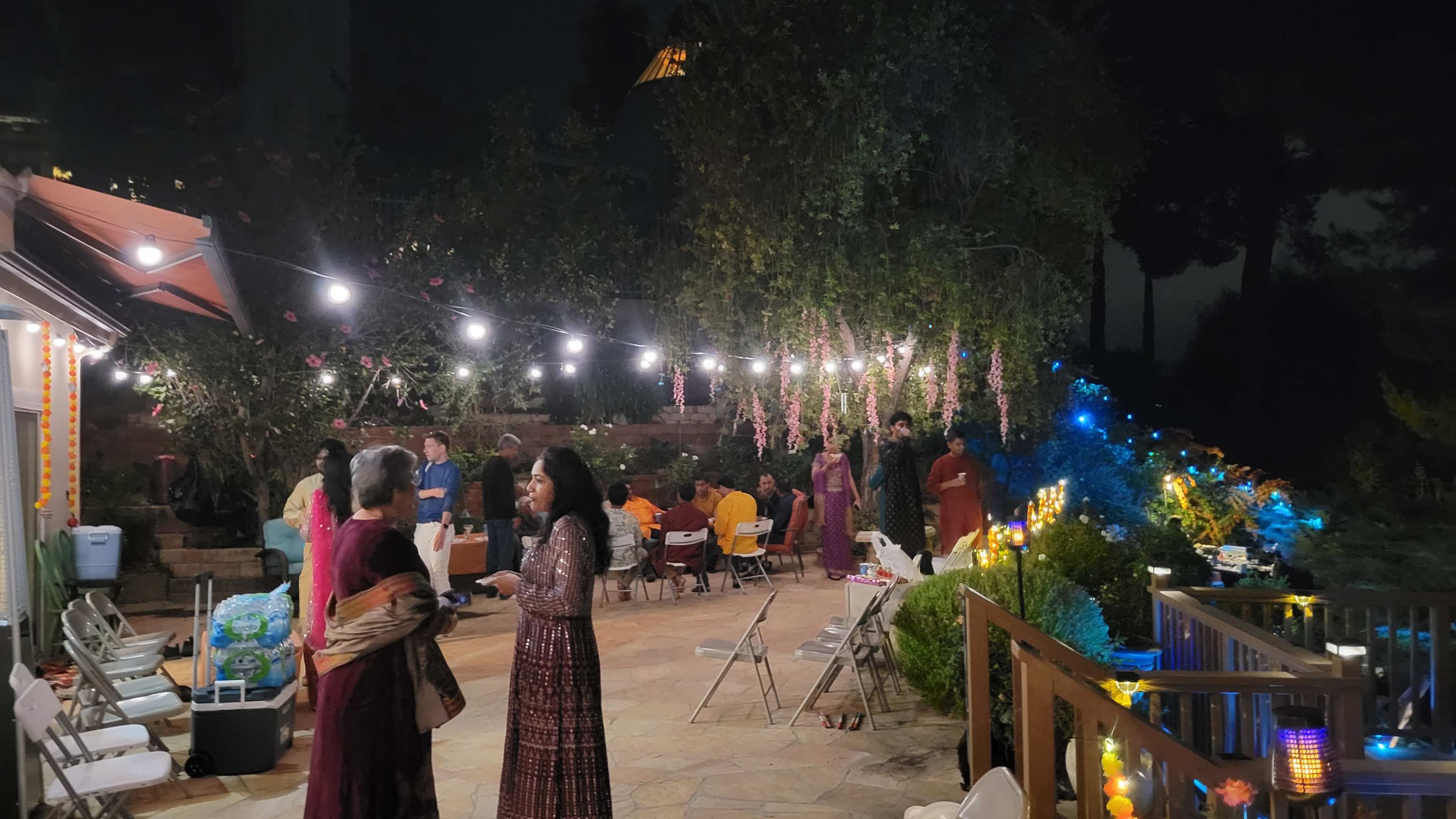 A group of people are gathered outdoors at night, surrounded by lights and decorations, with some seated at tables in the background.