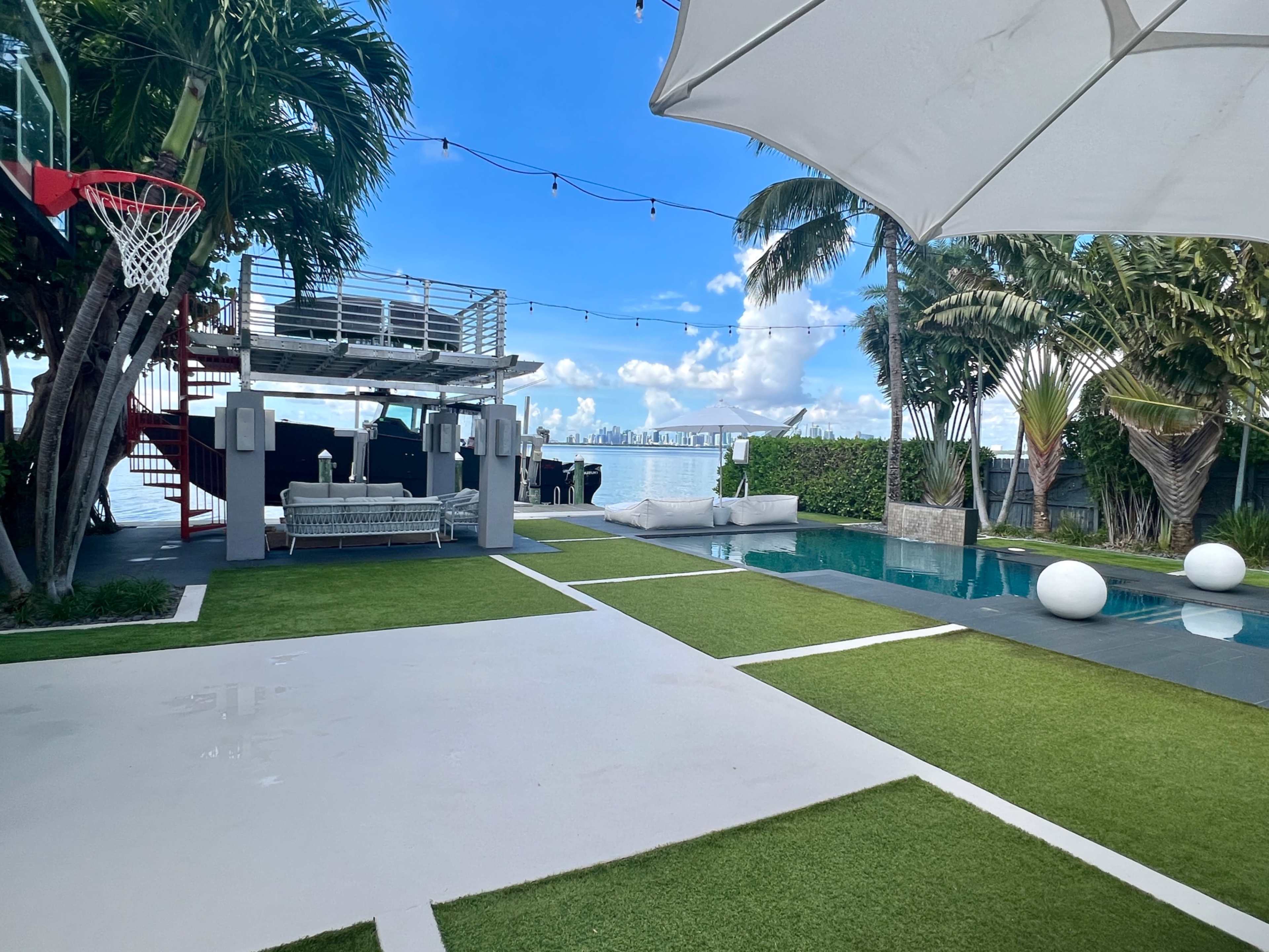 A modern outdoor space featuring a swimming pool, artificial turf, a basketball hoop, and a lounge area under a large white umbrella, with city skyline visible in the background.