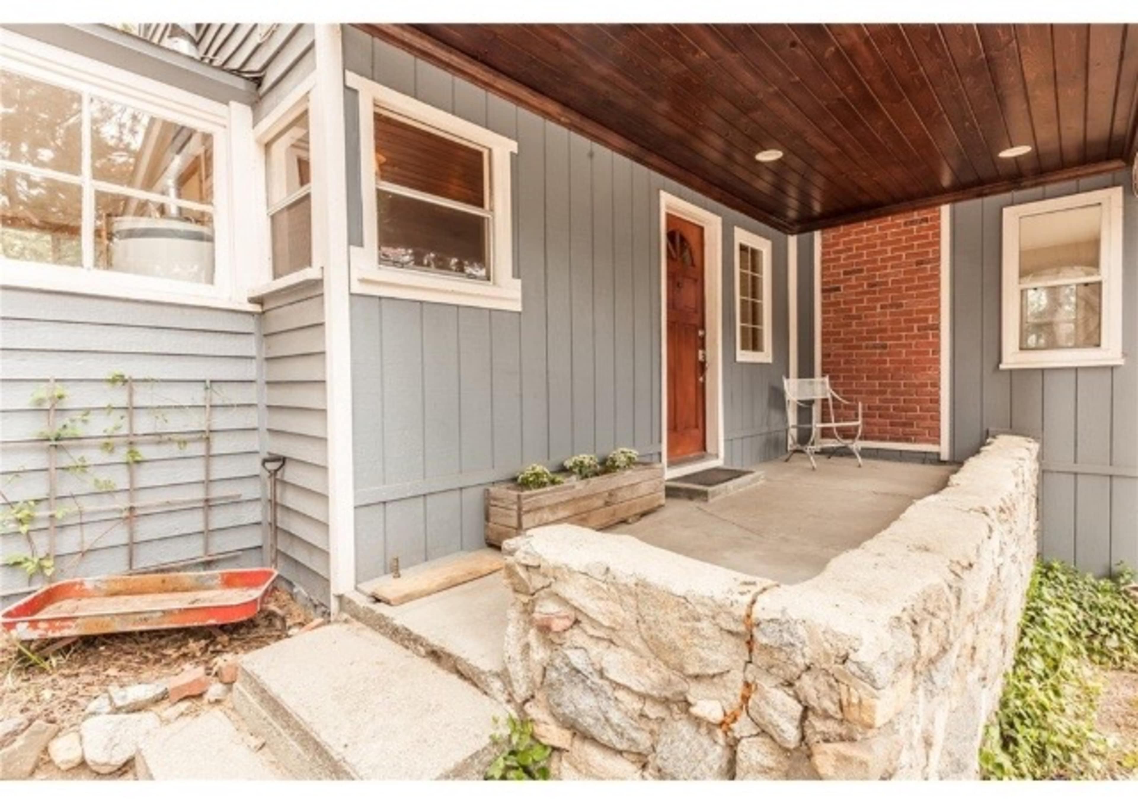 The image shows the entrance of a house with a stone pathway leading to a wooden door, surrounded by blue-gray siding and a covered porch area.