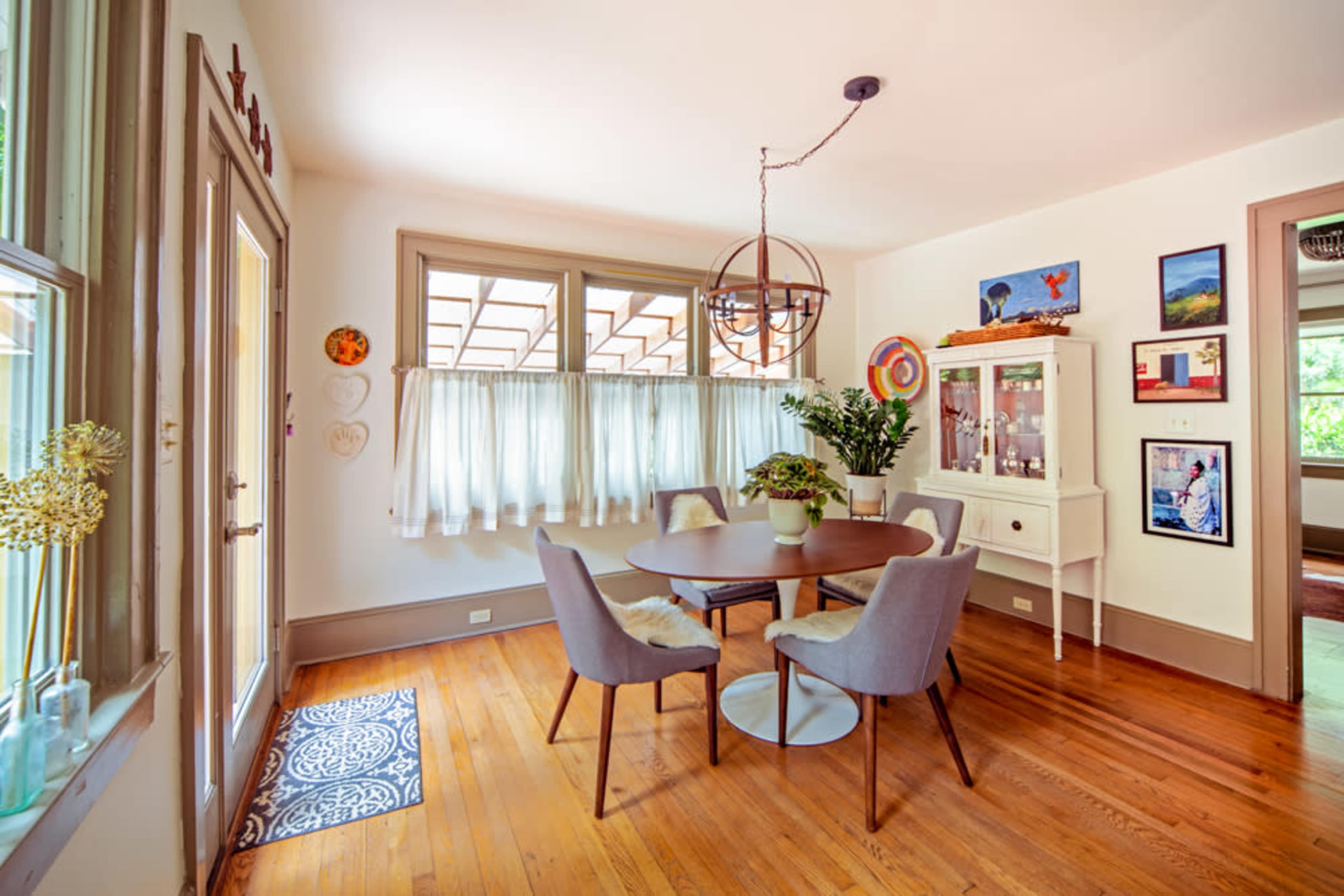 A well-lit dining area features a round table surrounded by four chairs, with large windows and decorative wall art.