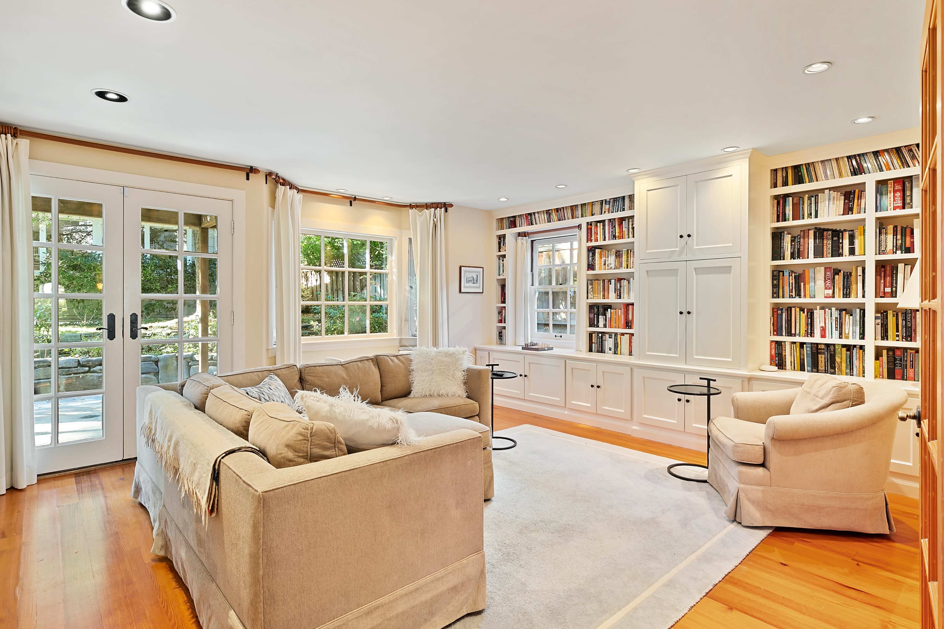 A bright living room features a beige sectional sofa, a pair of small side tables, and built-in bookshelves filled with books, along with French doors that open to a garden view.