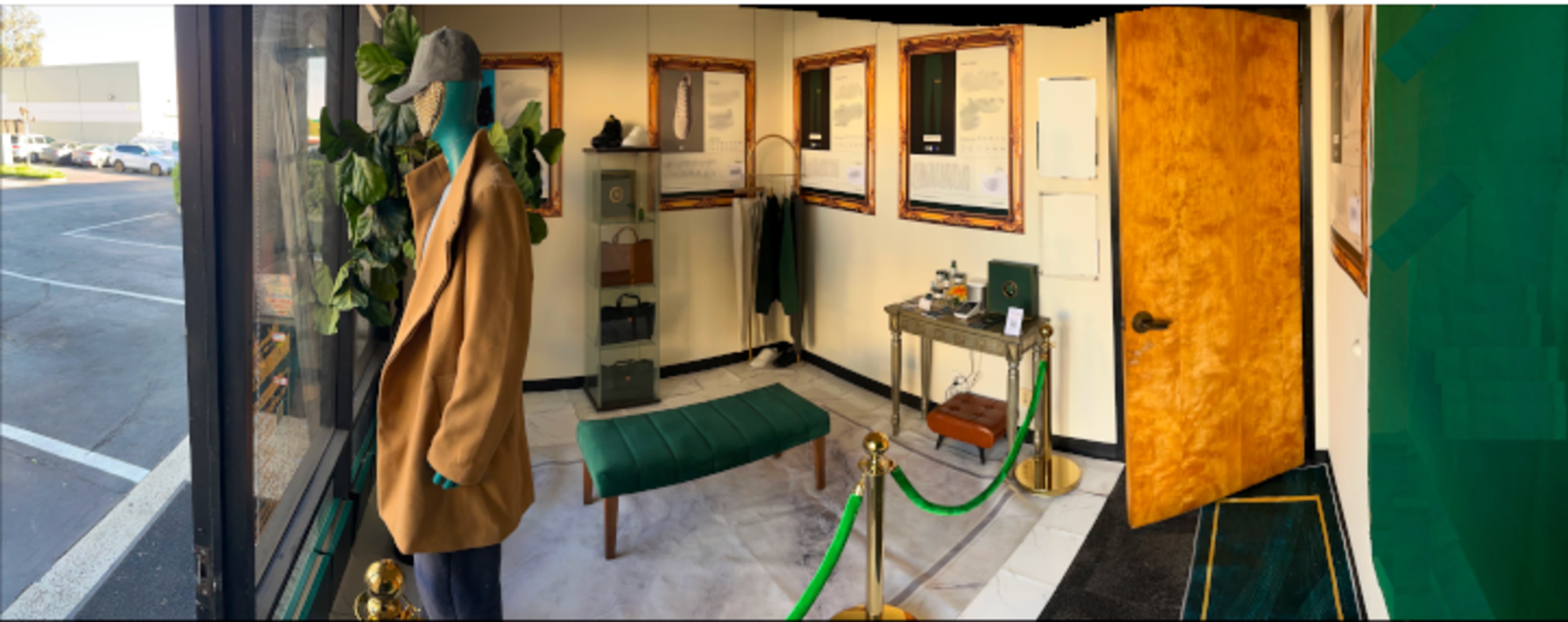 The image shows a display room featuring a seated mannequin dressed in a brown coat beside a green bench, with a small table and various items displayed on the walls.