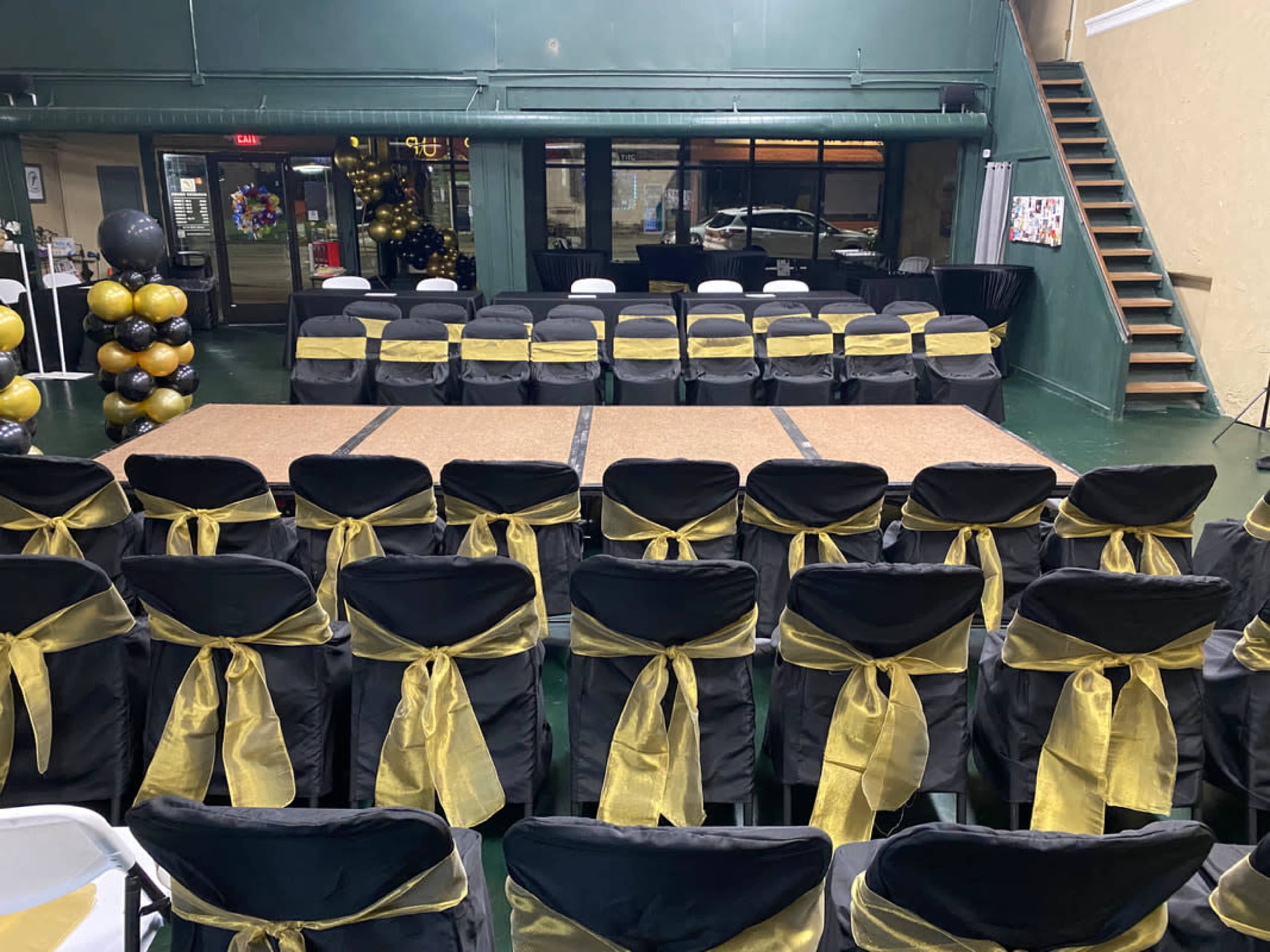 A spacious event venue set up with rows of black chairs adorned with gold sashes, facing a central stage area with a wooden platform.