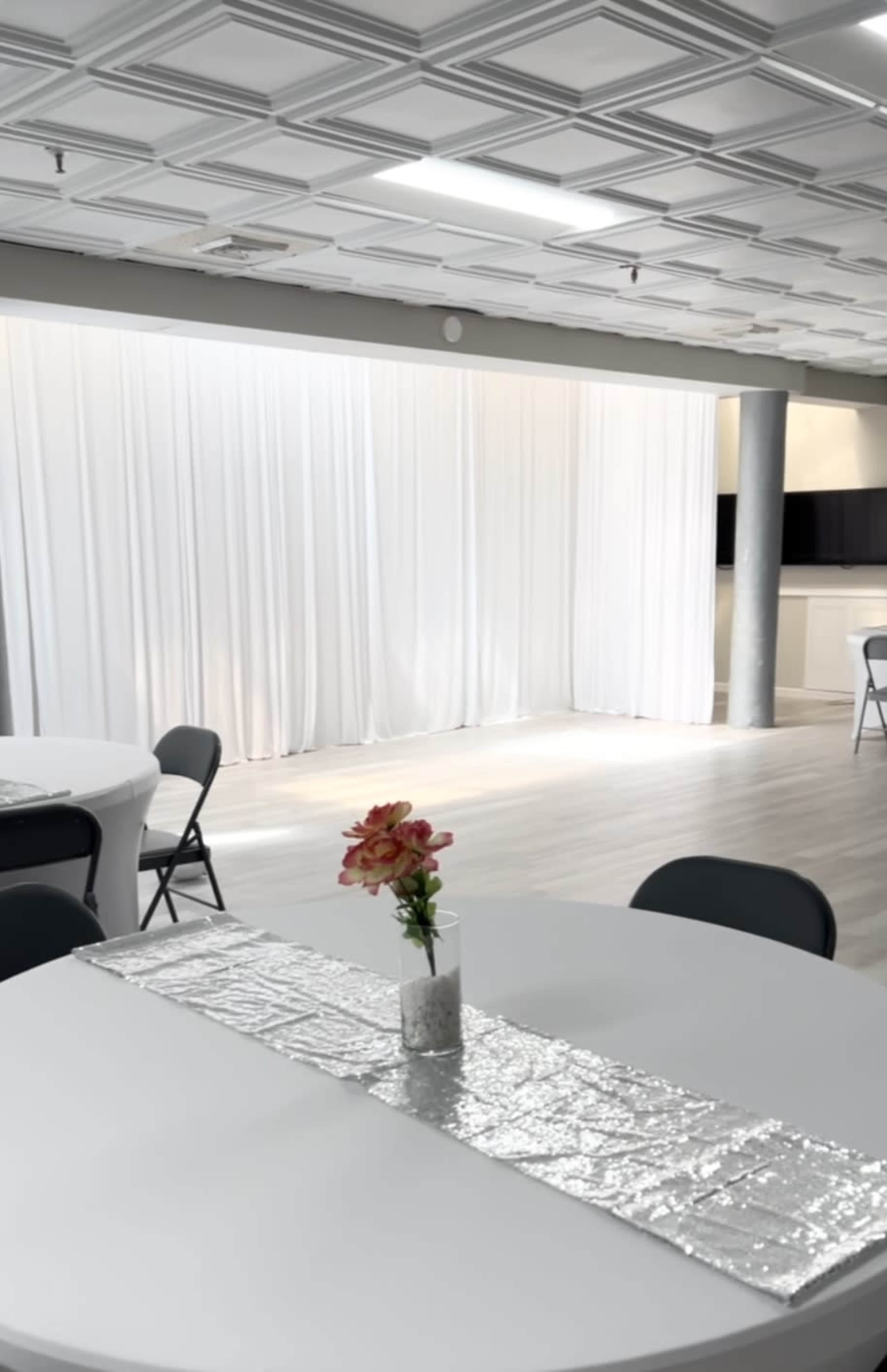 A bright event space features white drapes, circular tables with silver table runners, and a flower centerpiece.