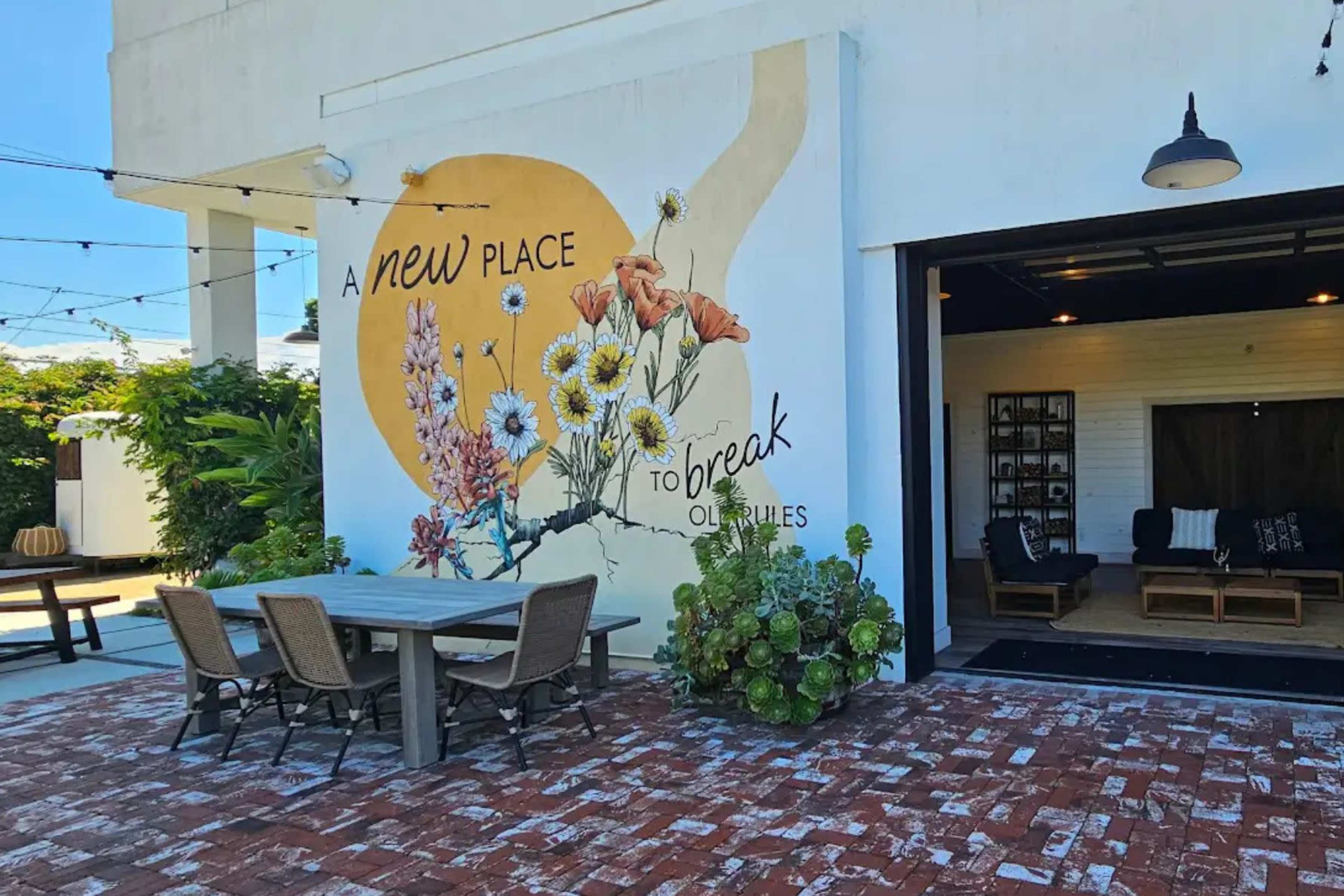 The image shows a patio area with a large mural depicting flowers and the text "A new place to break old rules" on the wall of a building.