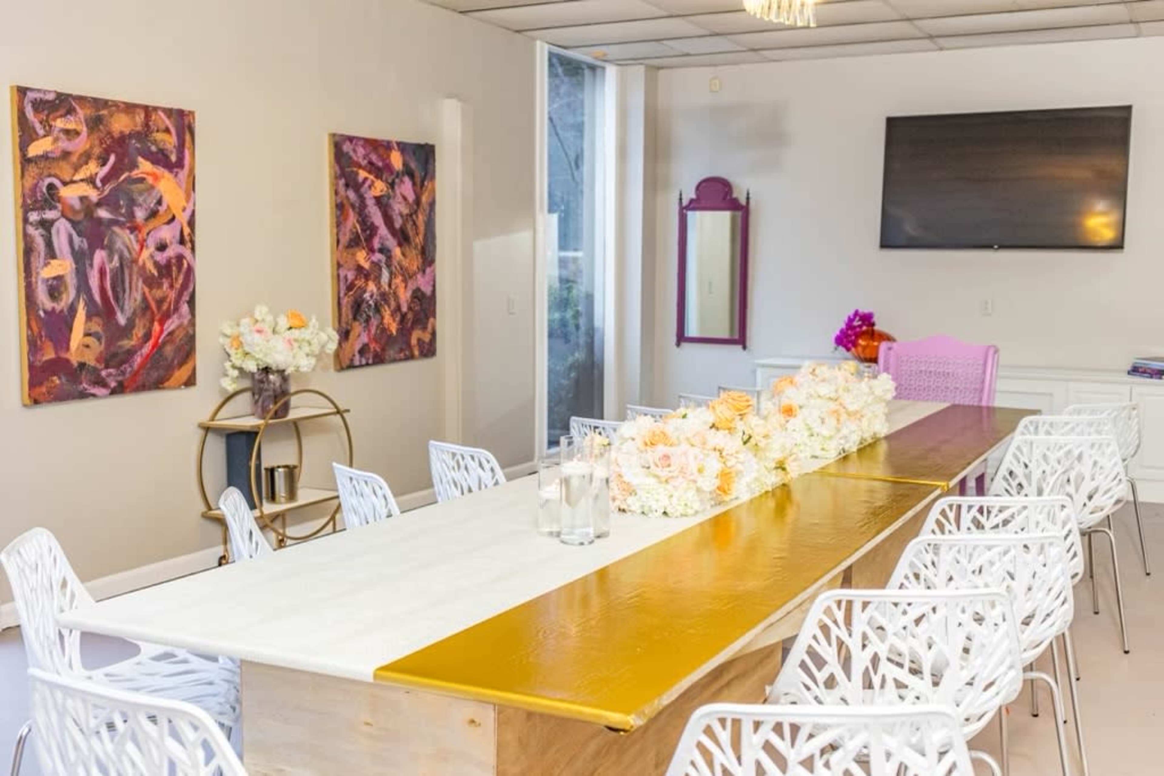 A dining area features a long table dressed in gold and white, surrounded by white chairs, with floral centerpieces and artwork on the walls.