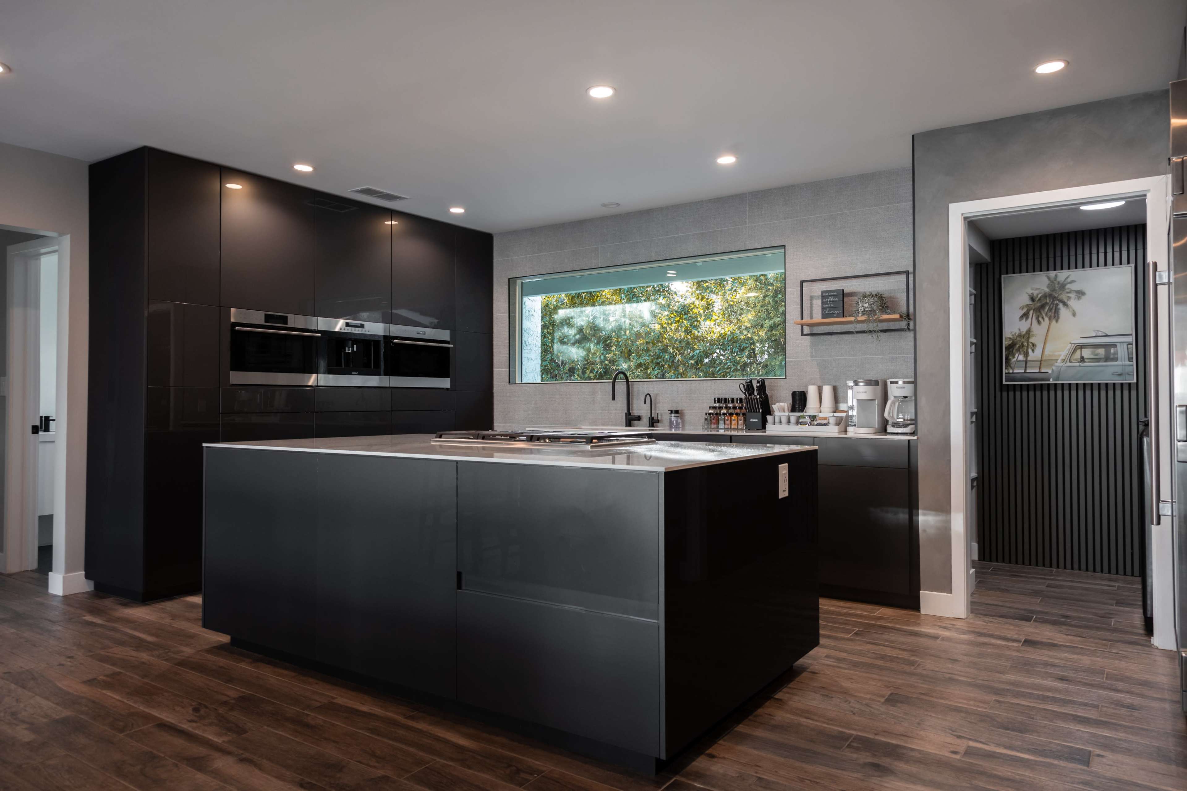 A modern kitchen features a large island with a sleek gray countertop, dark cabinetry, and a window overlooking greenery.