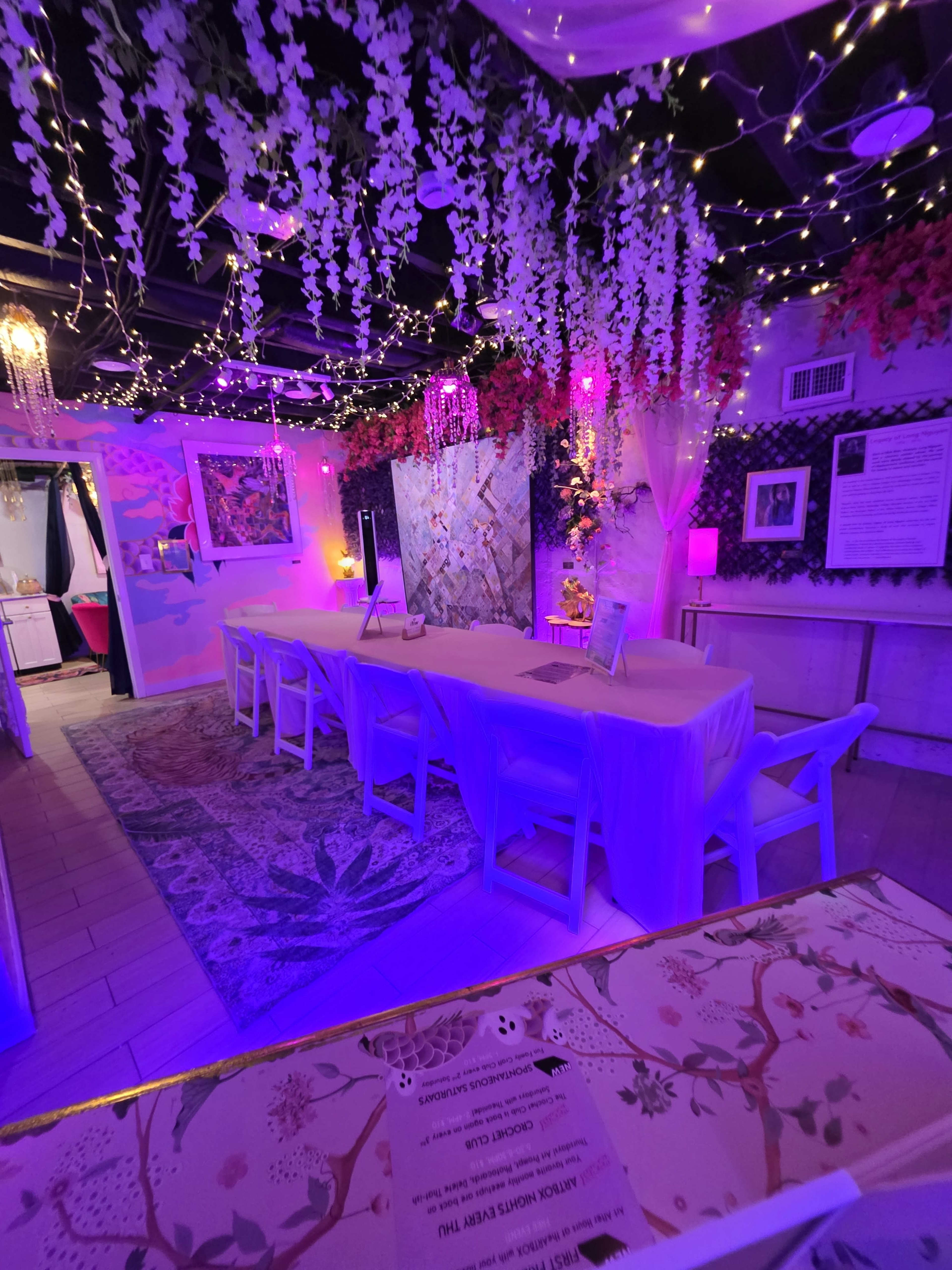 The image shows a decorated indoor space with a long white table, surrounded by colorful flowers and string lights.