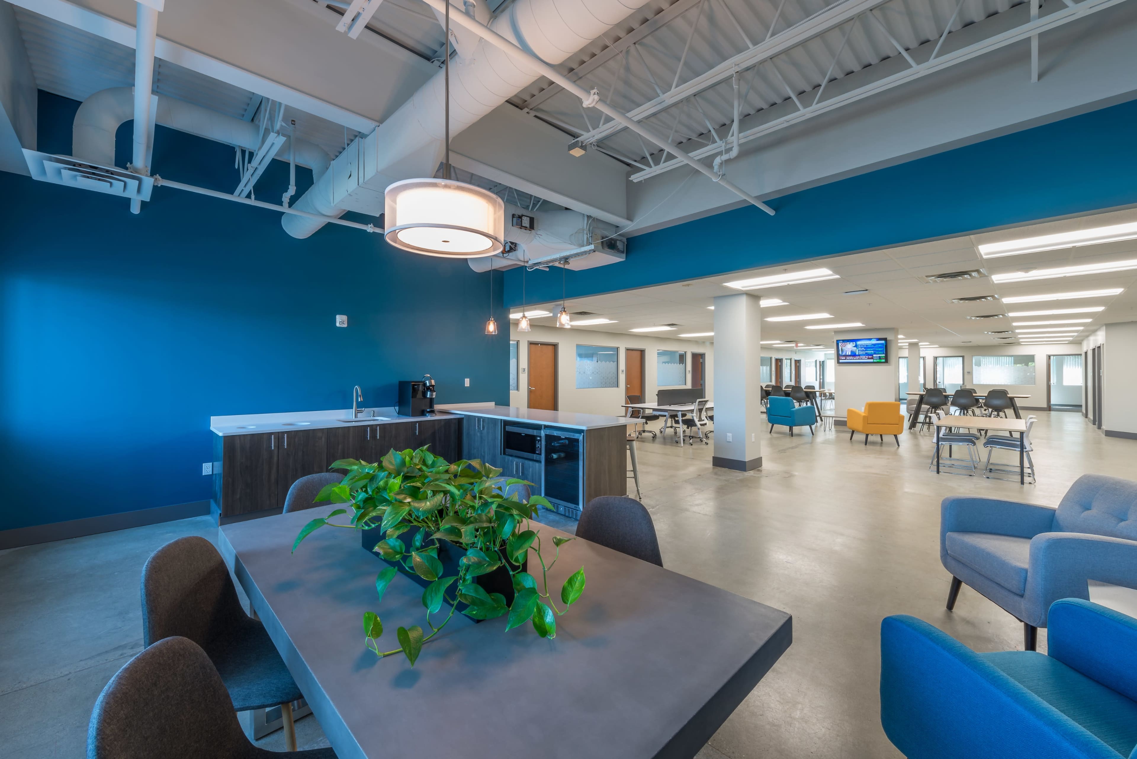 The image shows a modern office space featuring a kitchen area, a dining table with chairs, and multiple workstations with seating arrangements.