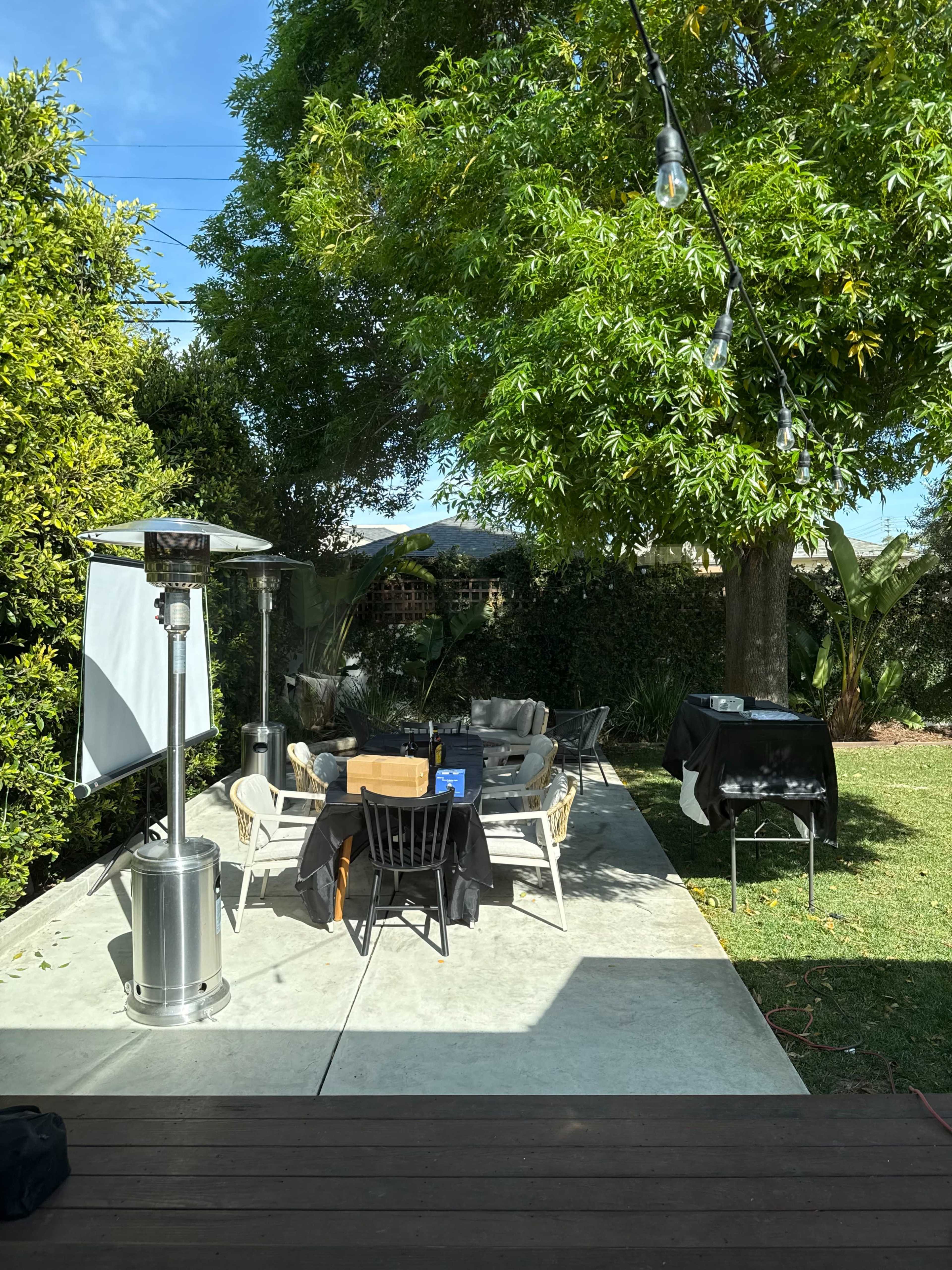 The image shows a backyard patio with seating arranged around a table, a patio heater, and a barbecue grill surrounded by greenery.