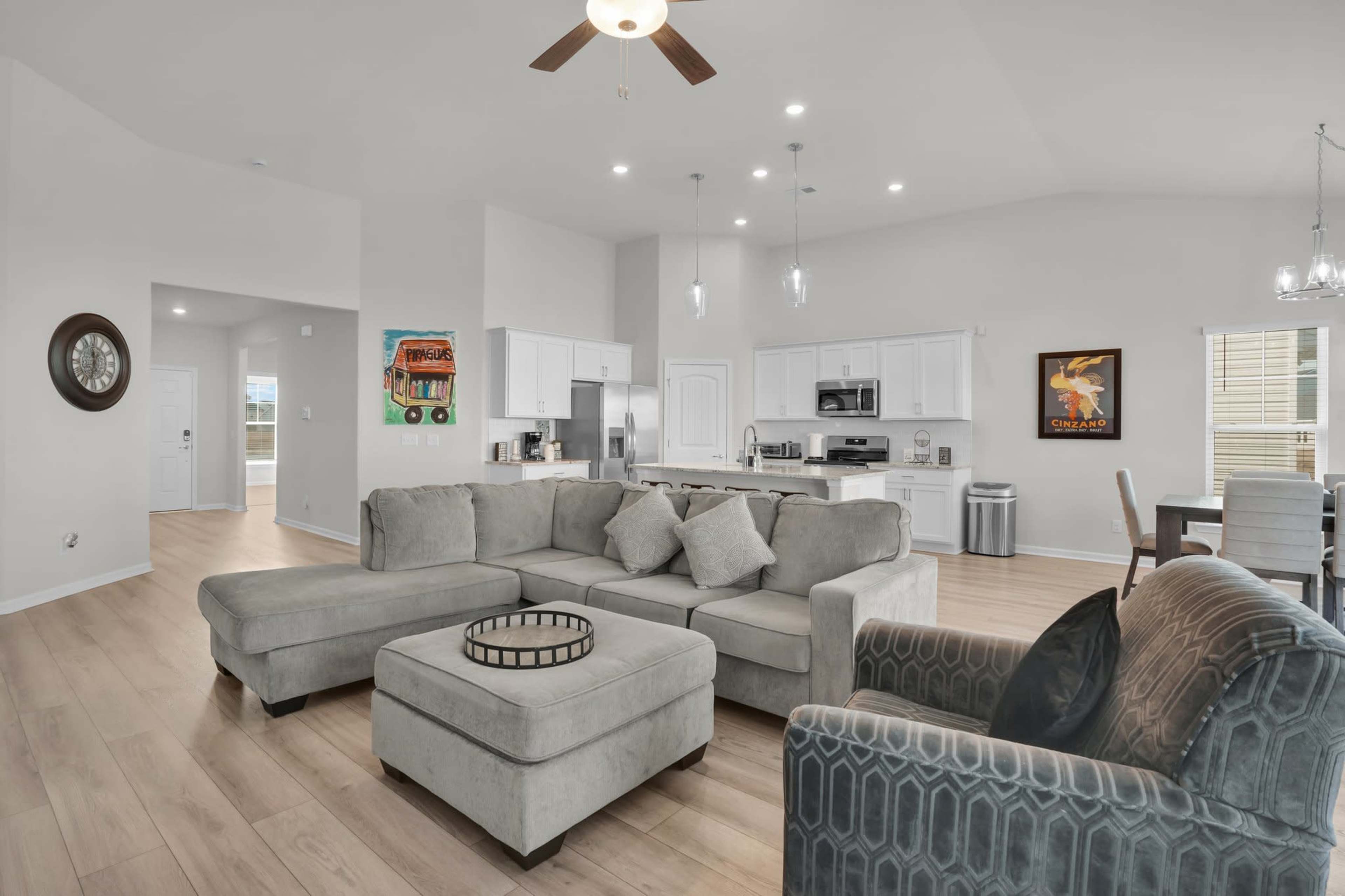 The image shows a modern living room with a sectional sofa, a coffee table, and a dining area, all set in an open-concept layout with light-colored walls and hardwood floors.