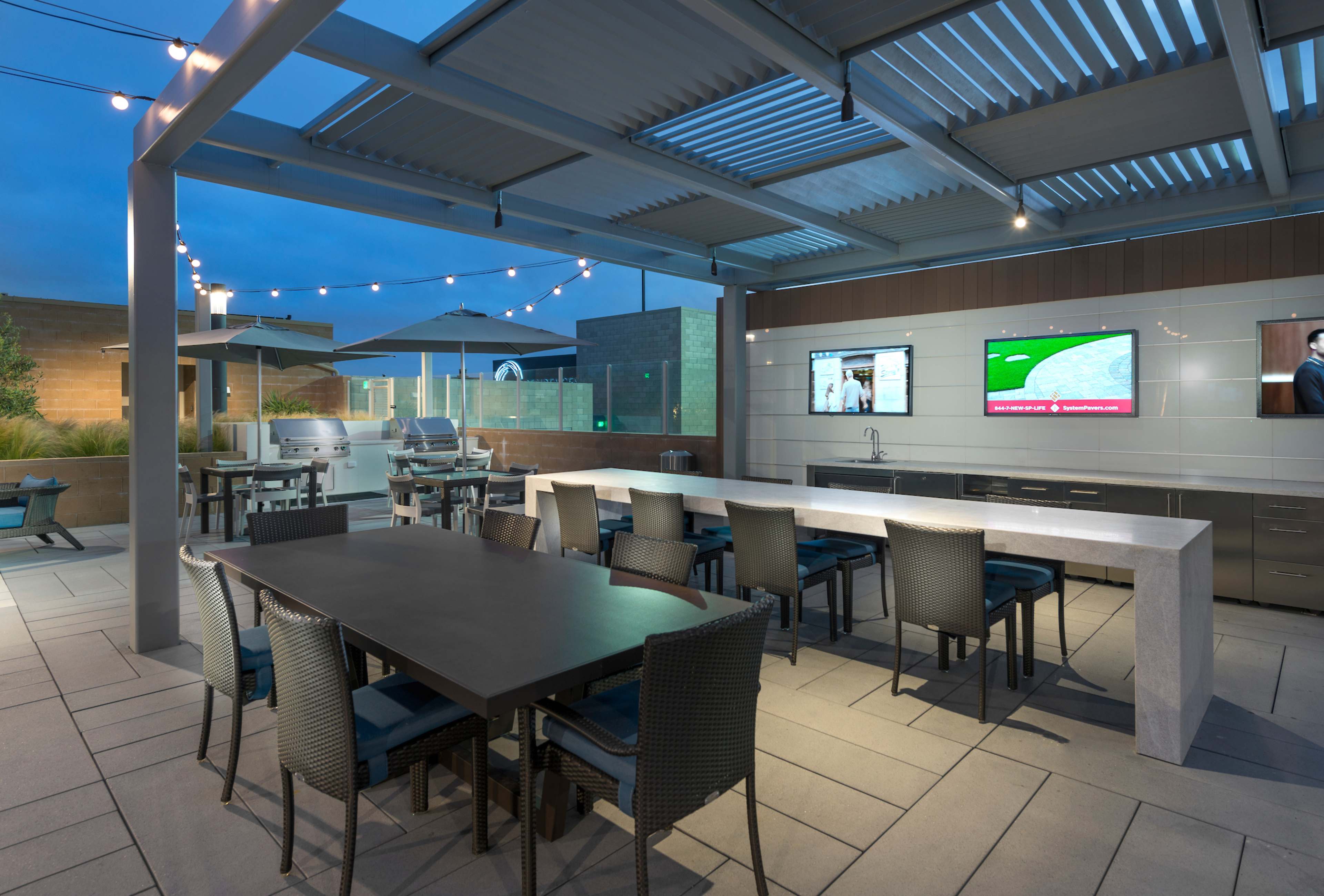 The image shows an outdoor dining area featuring a large table with chairs, an outdoor kitchen setup, and two televisions mounted on a wall under a canopy with string lights.