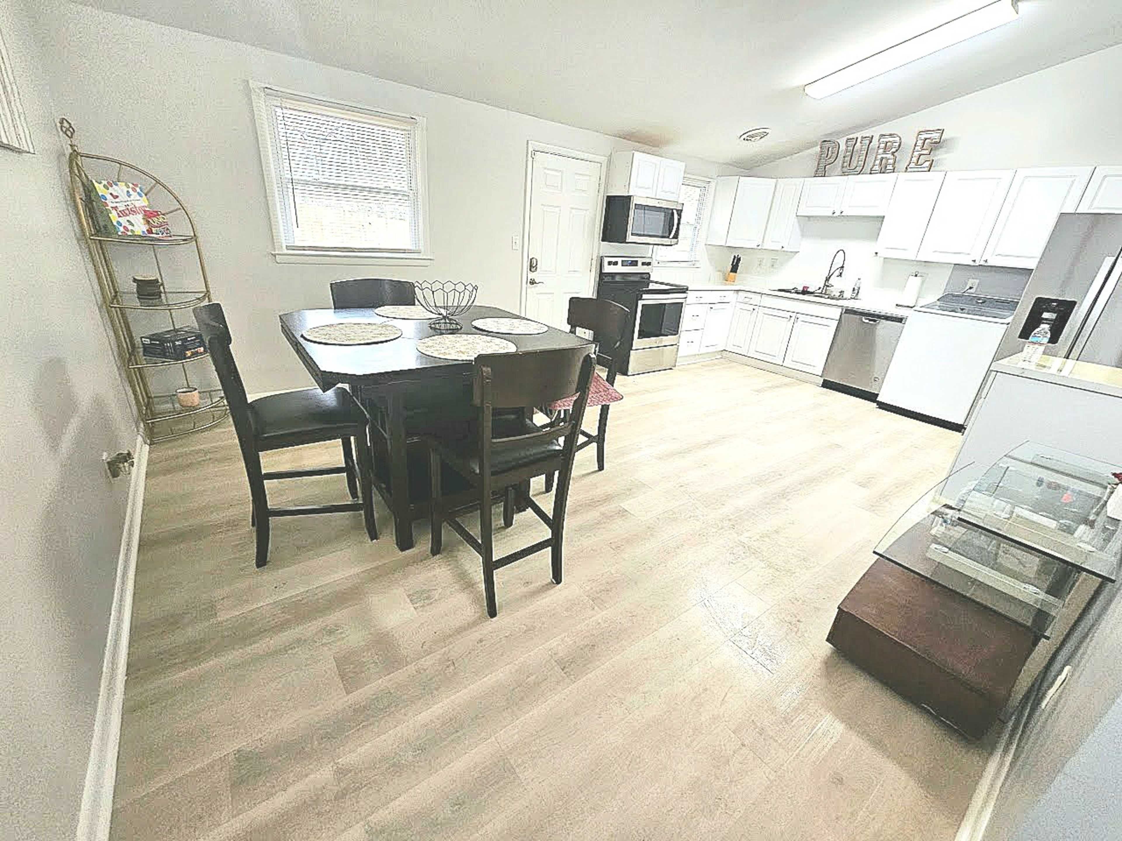 The image shows a modern kitchen and dining area with white cabinets, a dark wooden table set with chairs, and stainless steel appliances.